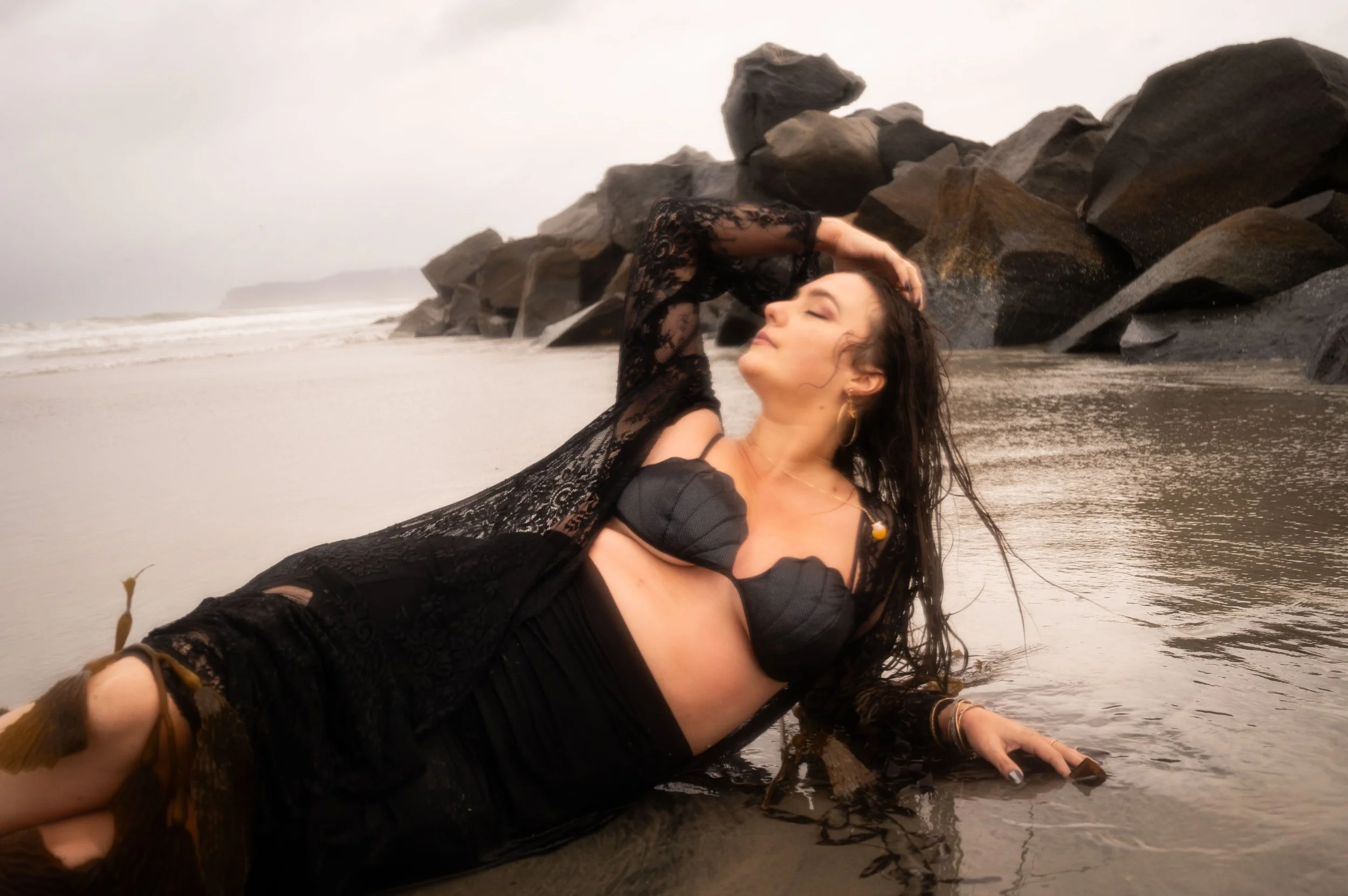Woman in black lace outfit reclining on the beach near rocks, with water and cloudy sky in background.