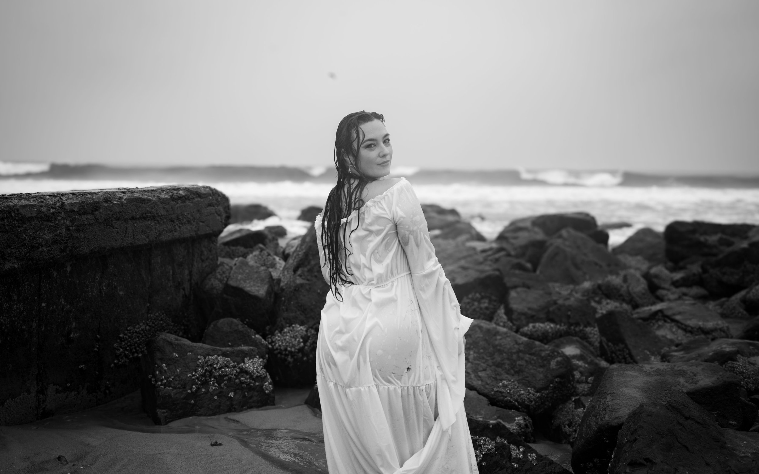 A woman wearing a white dress with wet hair, standing on rocks near the ocean, looking over her shoulder towards the camera, black and white photograph.