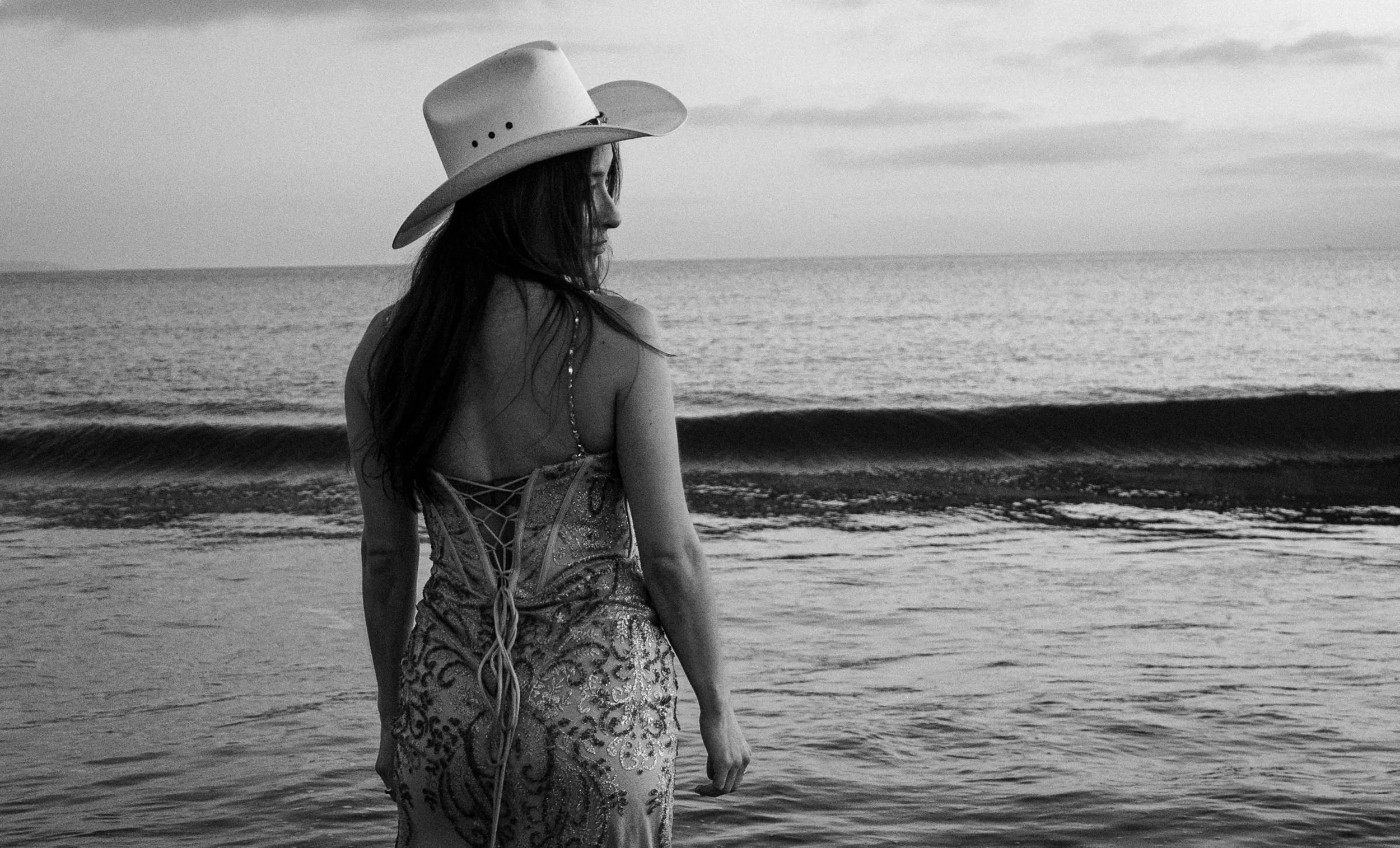 A woman with long hair wearing a cowboy hat and a detailed dress stands on the beach facing the sea in a black-and-white photograph.