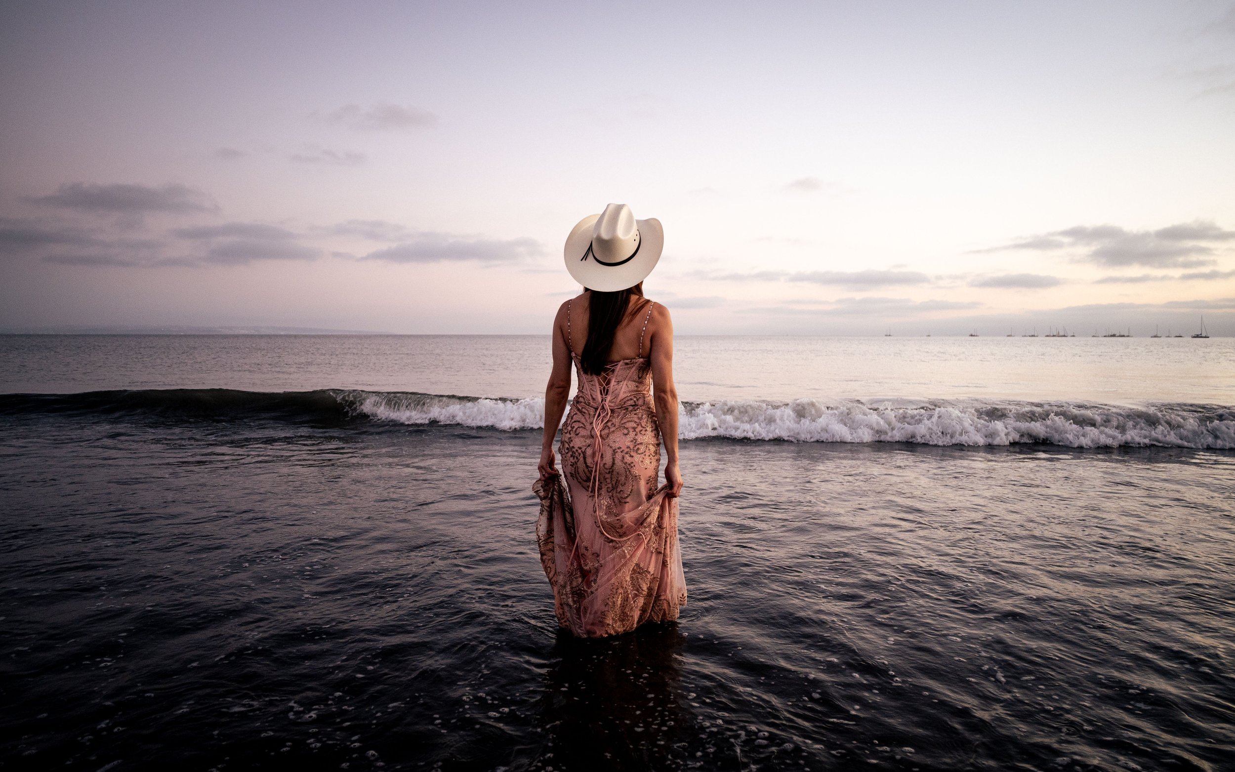 A woman in a pink dress and a wide-brimmed hat standing in shallow ocean water facing the horizon at sunset or sunrise.