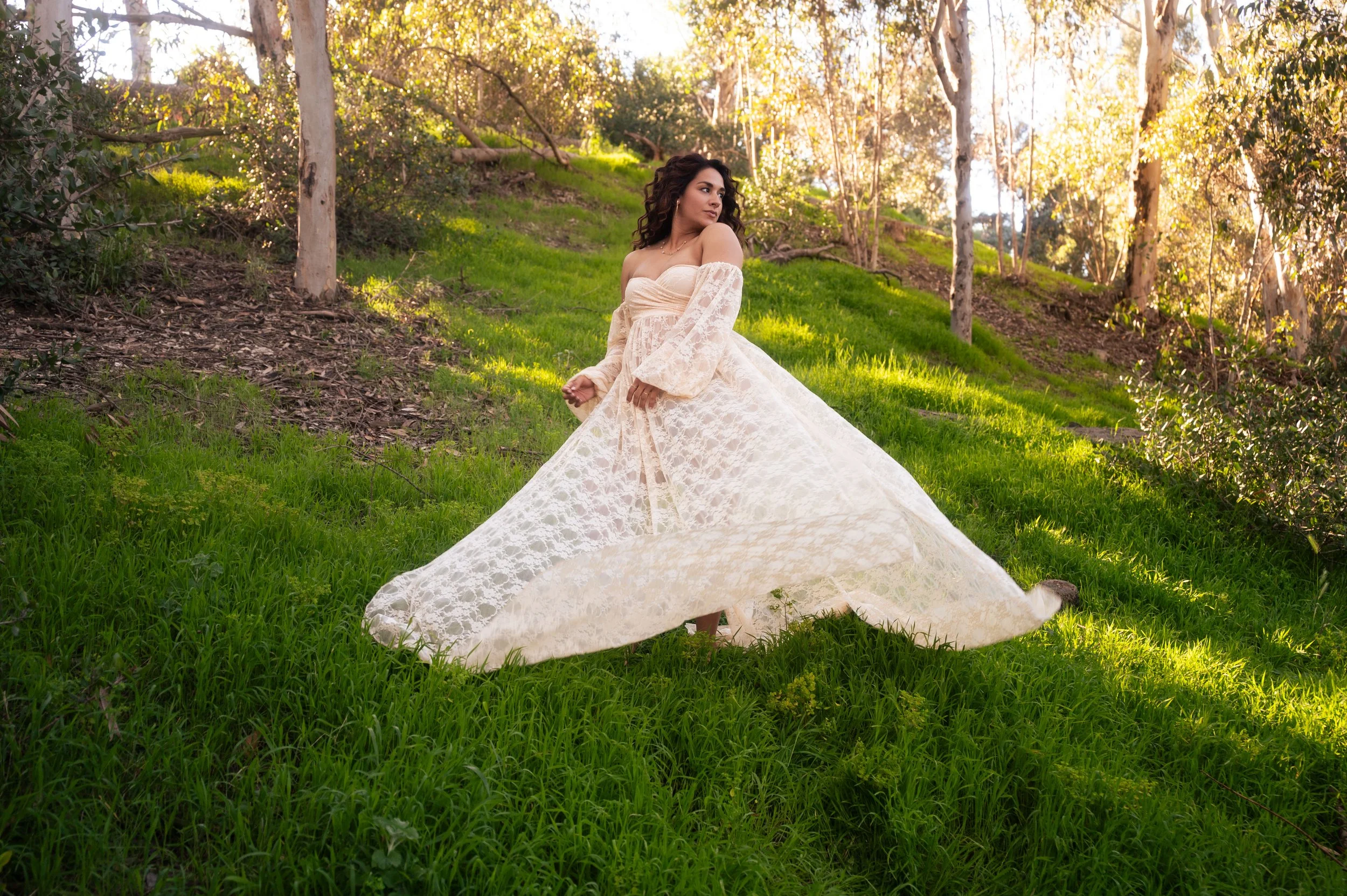 A woman in a flowing white lace dress standing outdoors on a grassy hillside surrounded by trees, with sunlight filtering through the leaves.