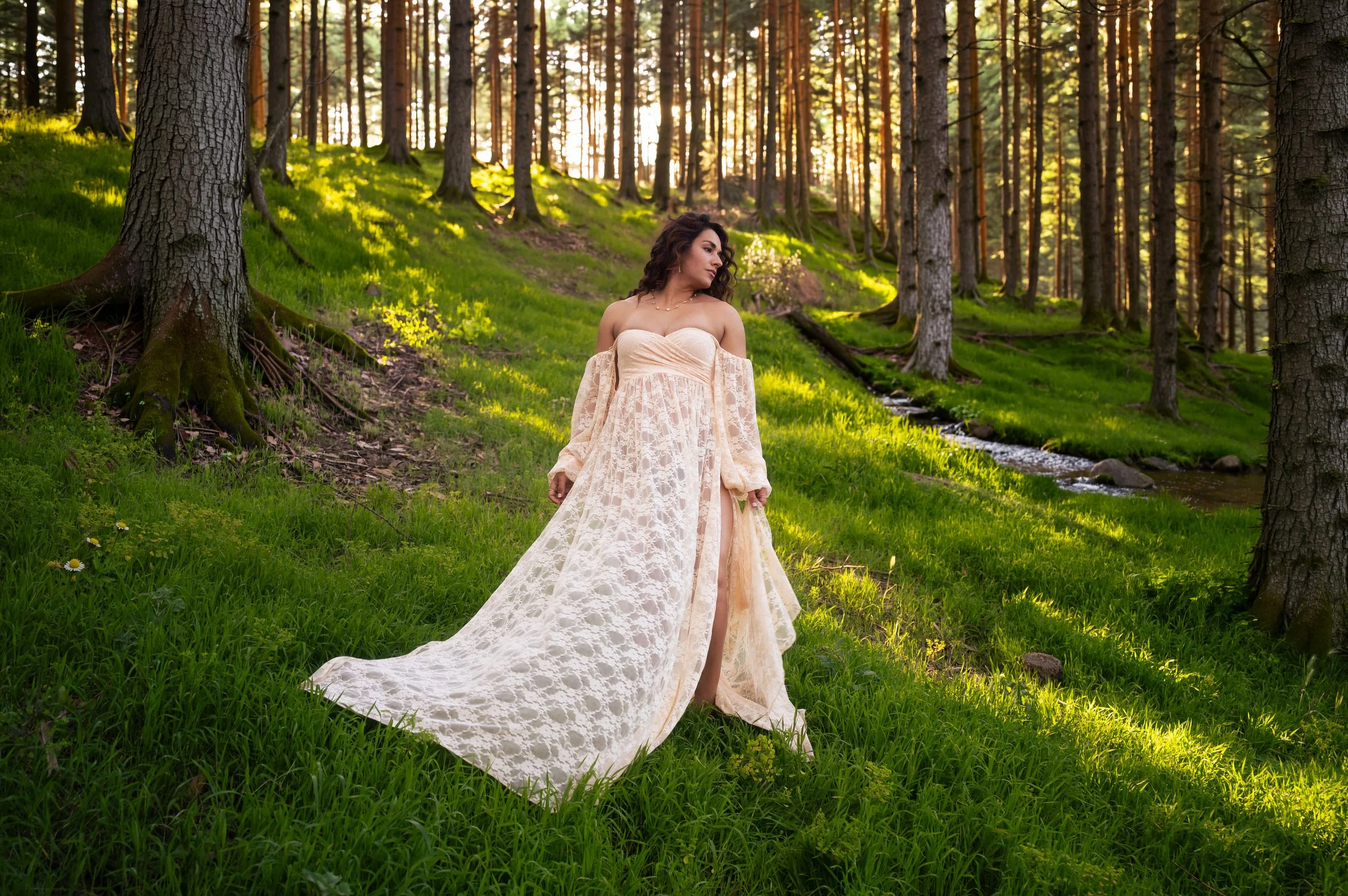 A woman in a flowing, off-the-shoulder lace dress stands in a sunlit forest with tall trees and green grass.
