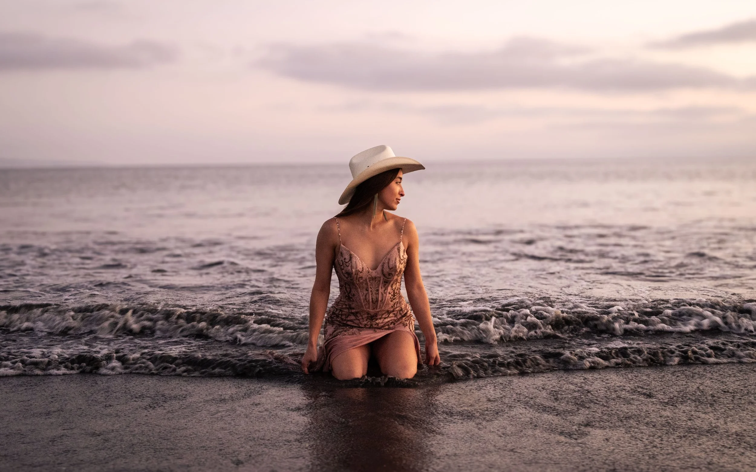 A woman wearing a wide-brimmed hat and a patterned dress sitting in shallow water at the beach during sunset.