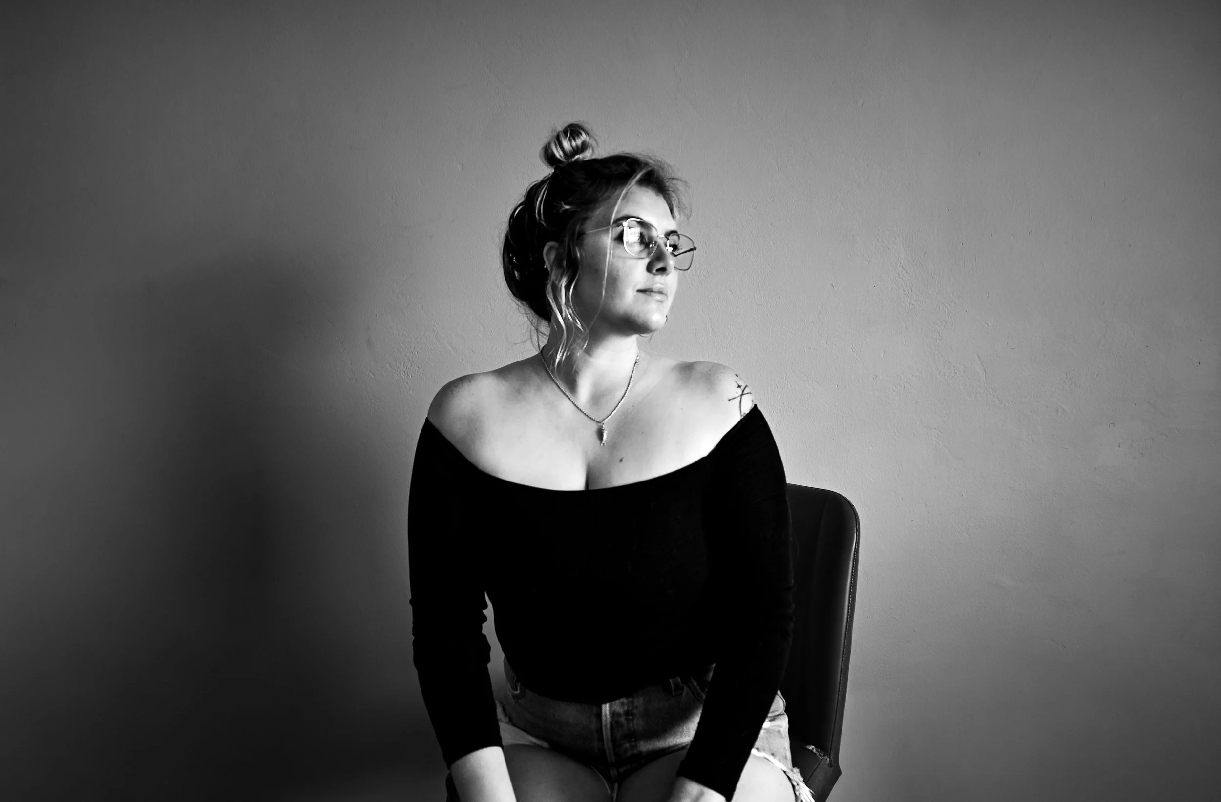 Black and white photo of a woman with glasses, a top bun hairstyle, wearing an off-the-shoulder top, sitting on a chair against a plain wall, looking away from the camera.