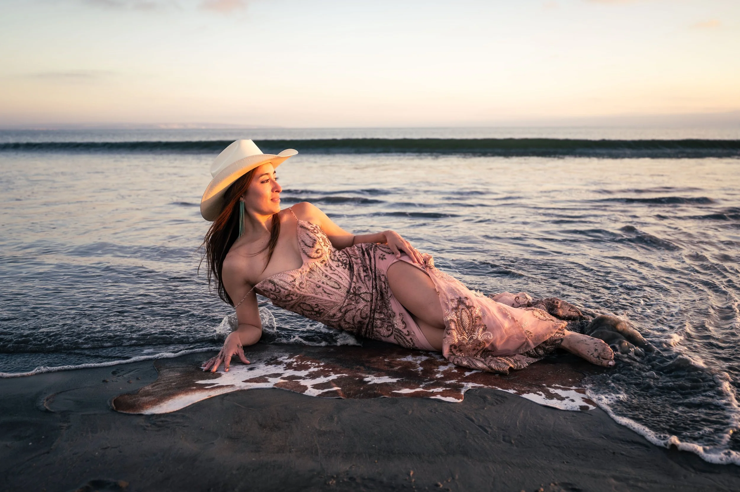 A woman in an elegant dress and cowboy hat reclining on the beach near the shoreline at sunset.