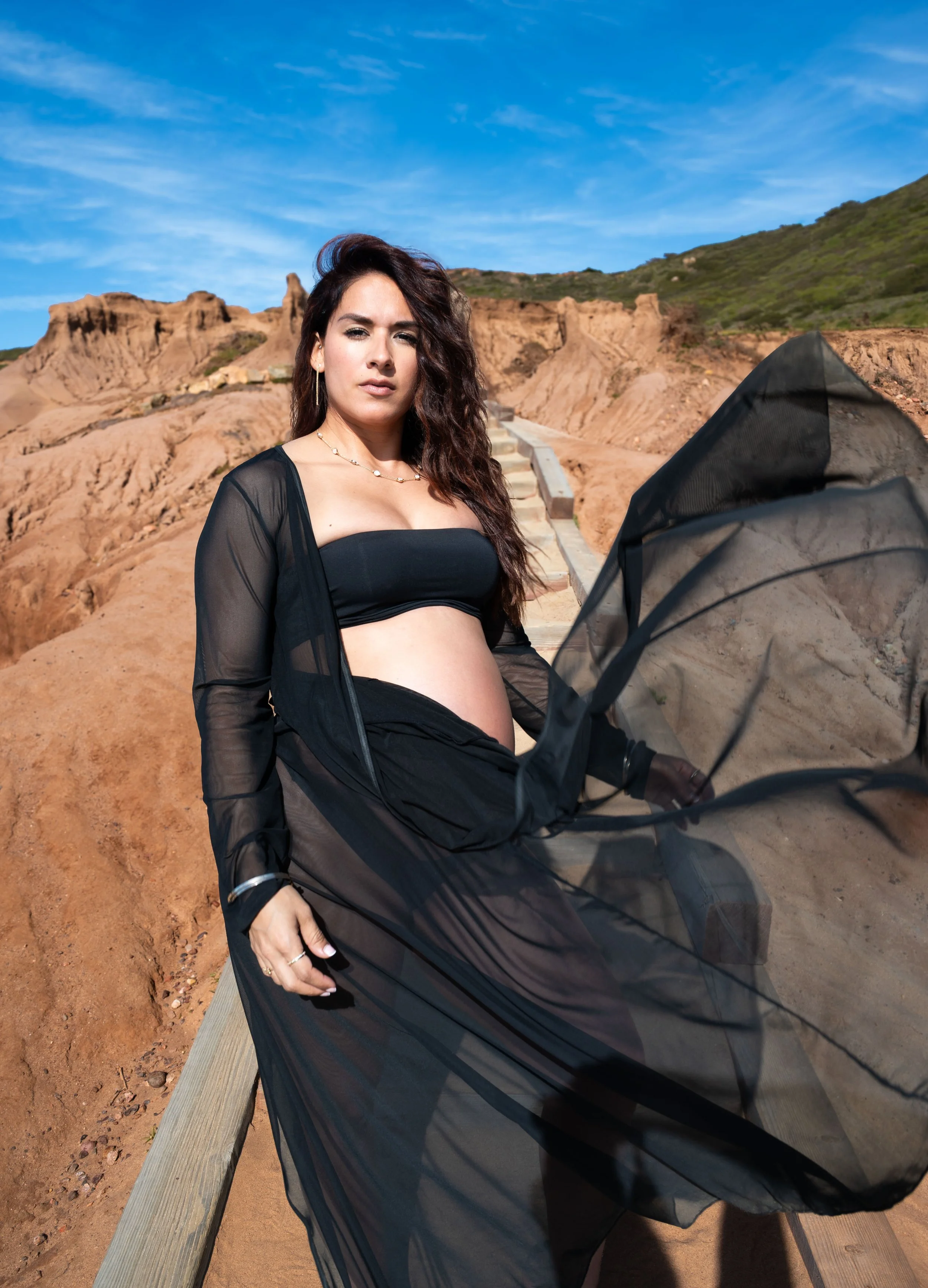 A pregnant woman wearing a black strapless top and sheer black cover-up sitting on a wooden bench outdoors, with a desert landscape and blue sky in the background.