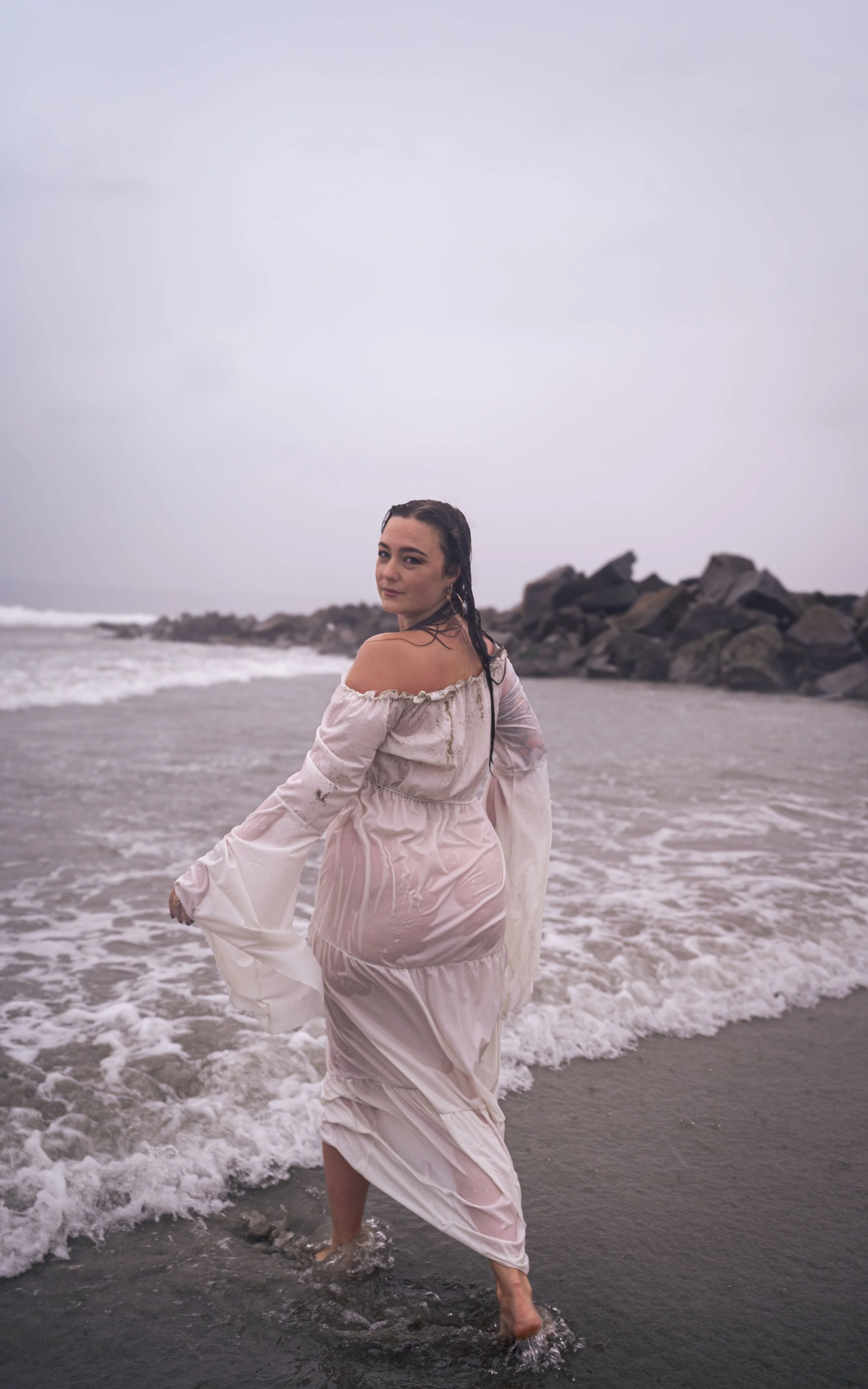 Woman in a wet, off-shoulder, light pink dress walking in the ocean waves with rocks in the background on a cloudy day.