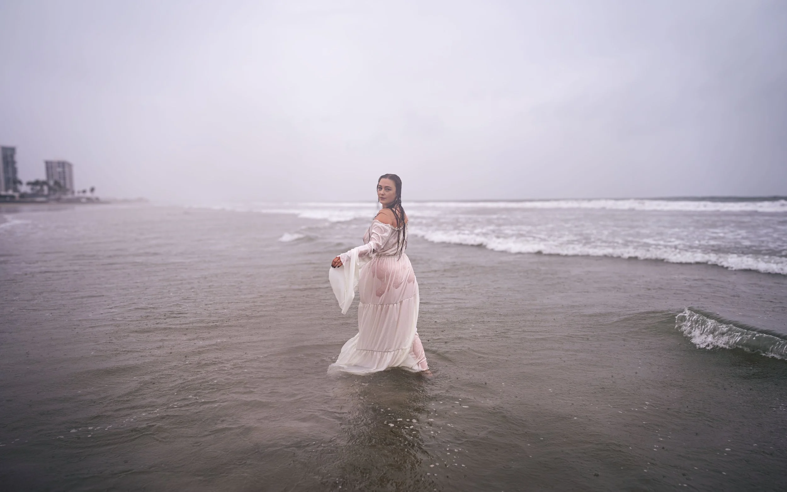 A woman in a white dress walking in shallow ocean water on a cloudy day