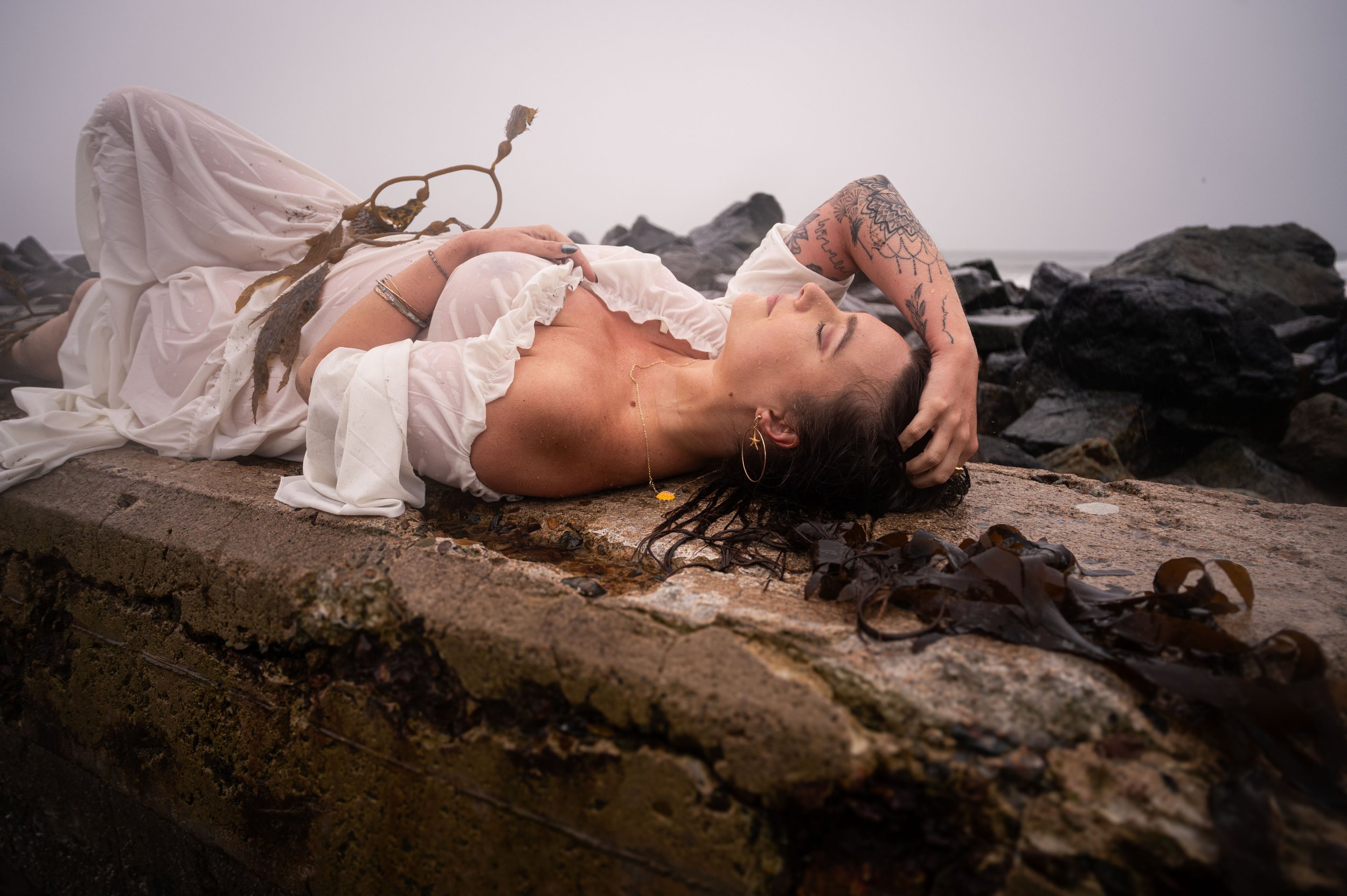 A woman lying on a rocky surface by the water, wearing a white dress, with her eyes closed and her left arm resting on her forehead.