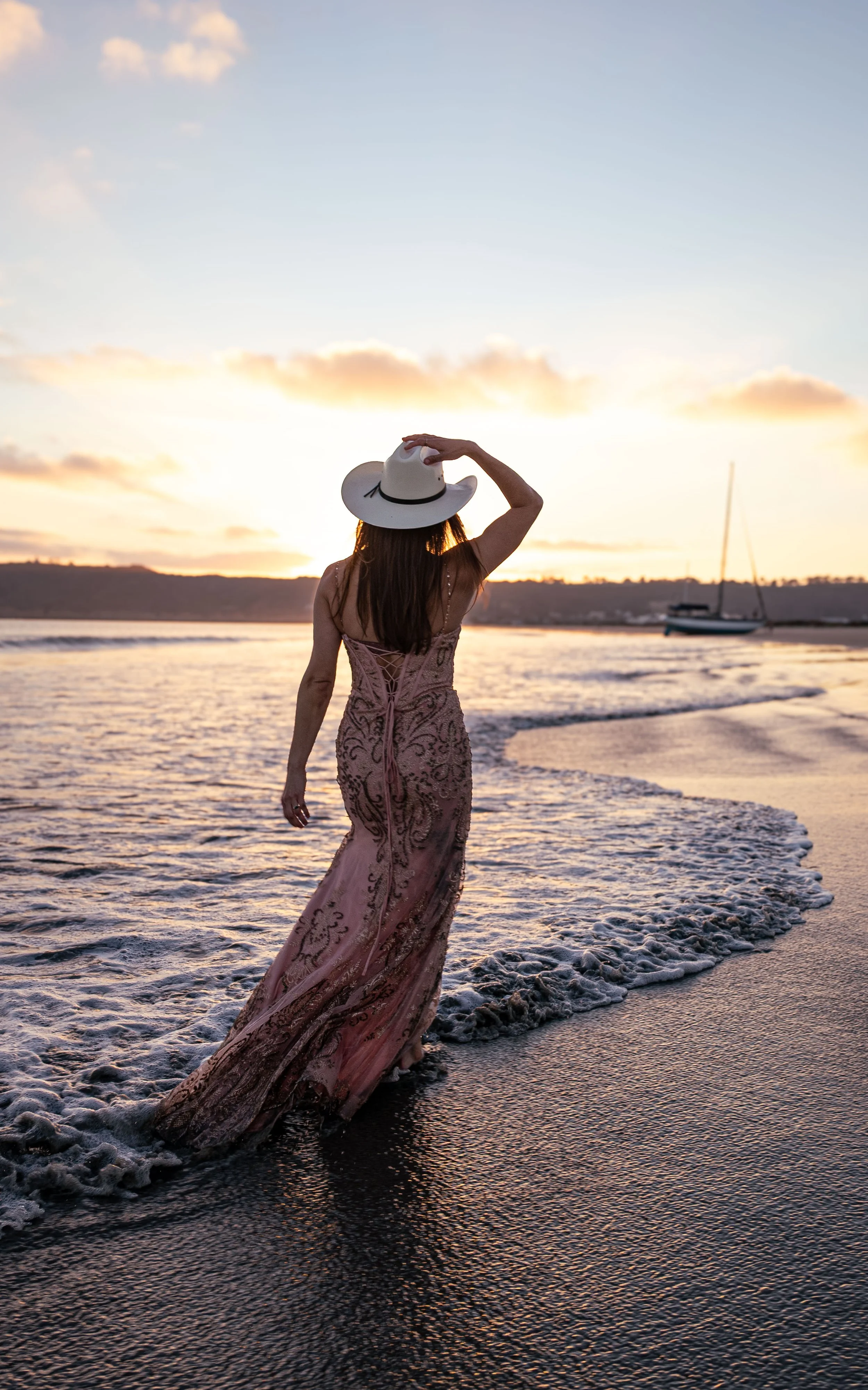 A woman in a long, elegant dress and a wide-brimmed hat walks along the shoreline at sunset, with a sailboat in the distance on the water.