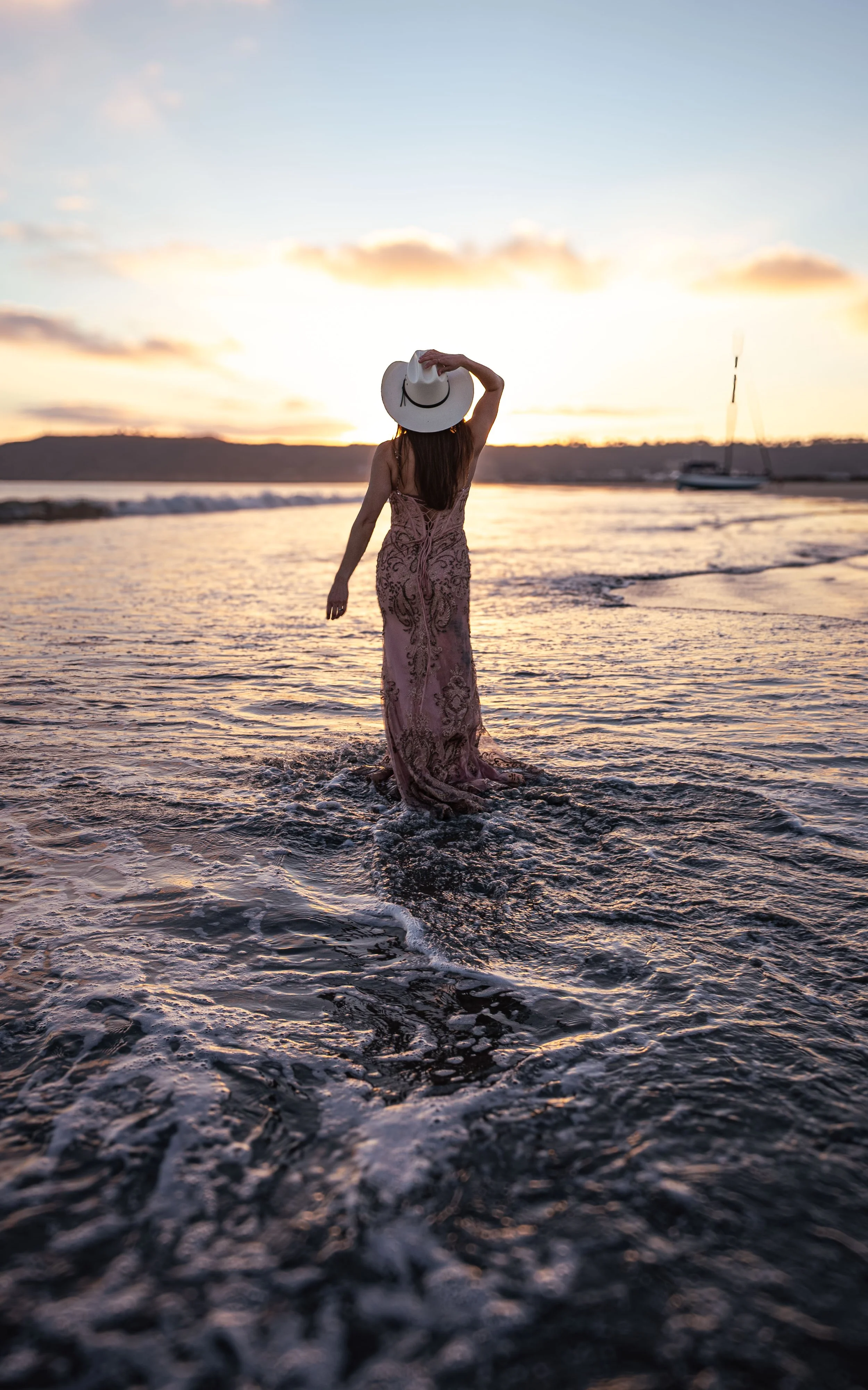 Woman in a long dress and wide-brimmed hat walking in shallow ocean water at sunset.