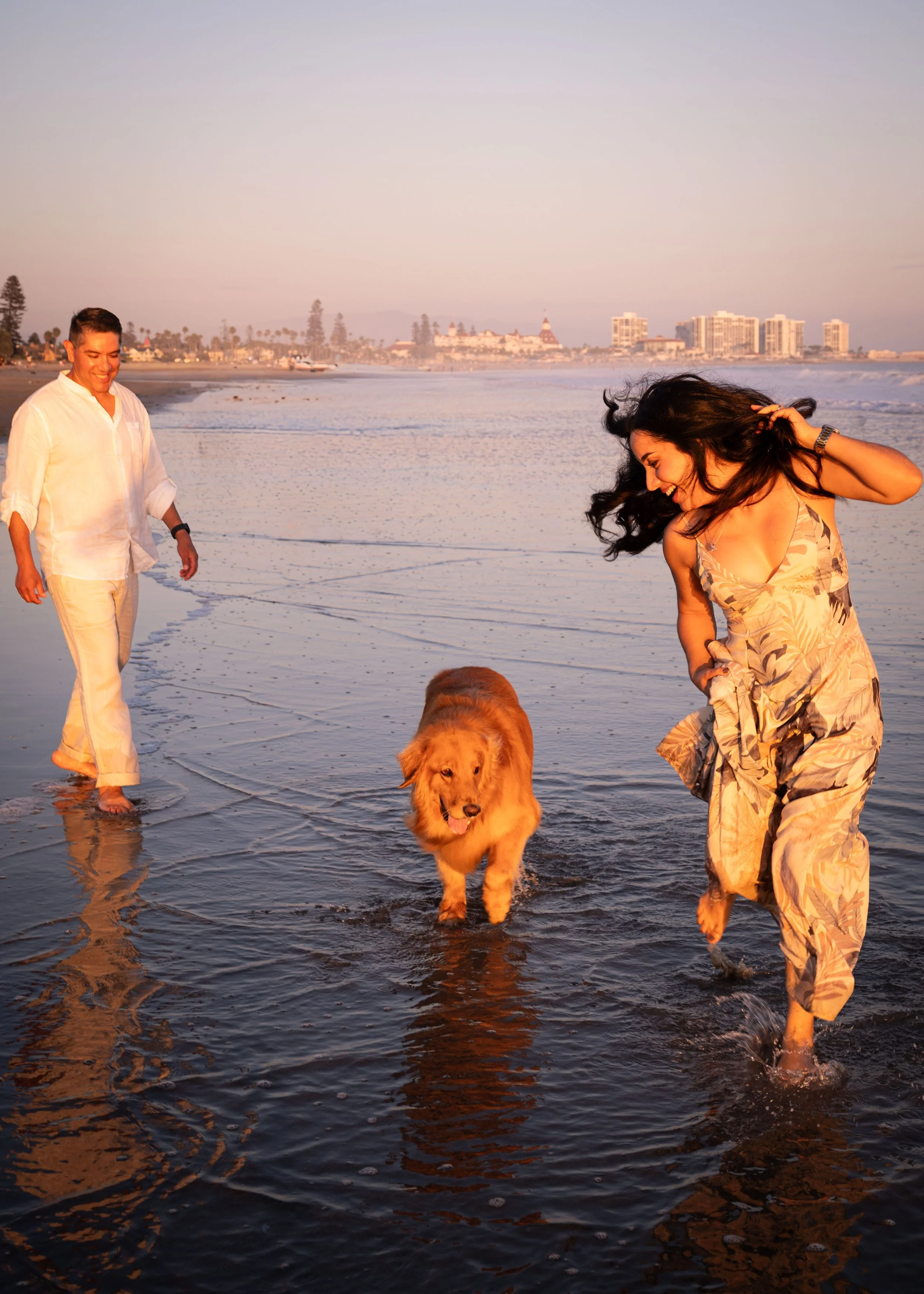 Two people walking a golden retriever dog on the beach at sunset, with city buildings in the background.