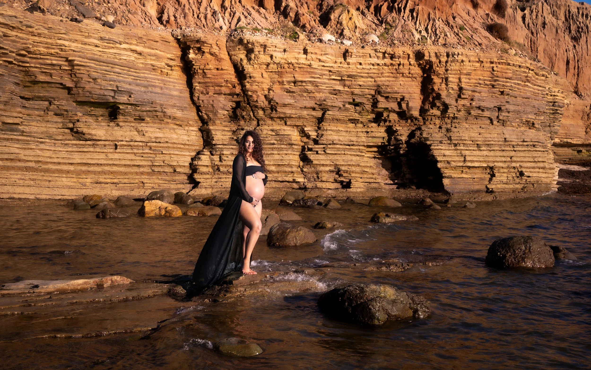 Pregnant woman in a black dress standing in shallow water near rocky beach with large rock formations and cliffs in the background.