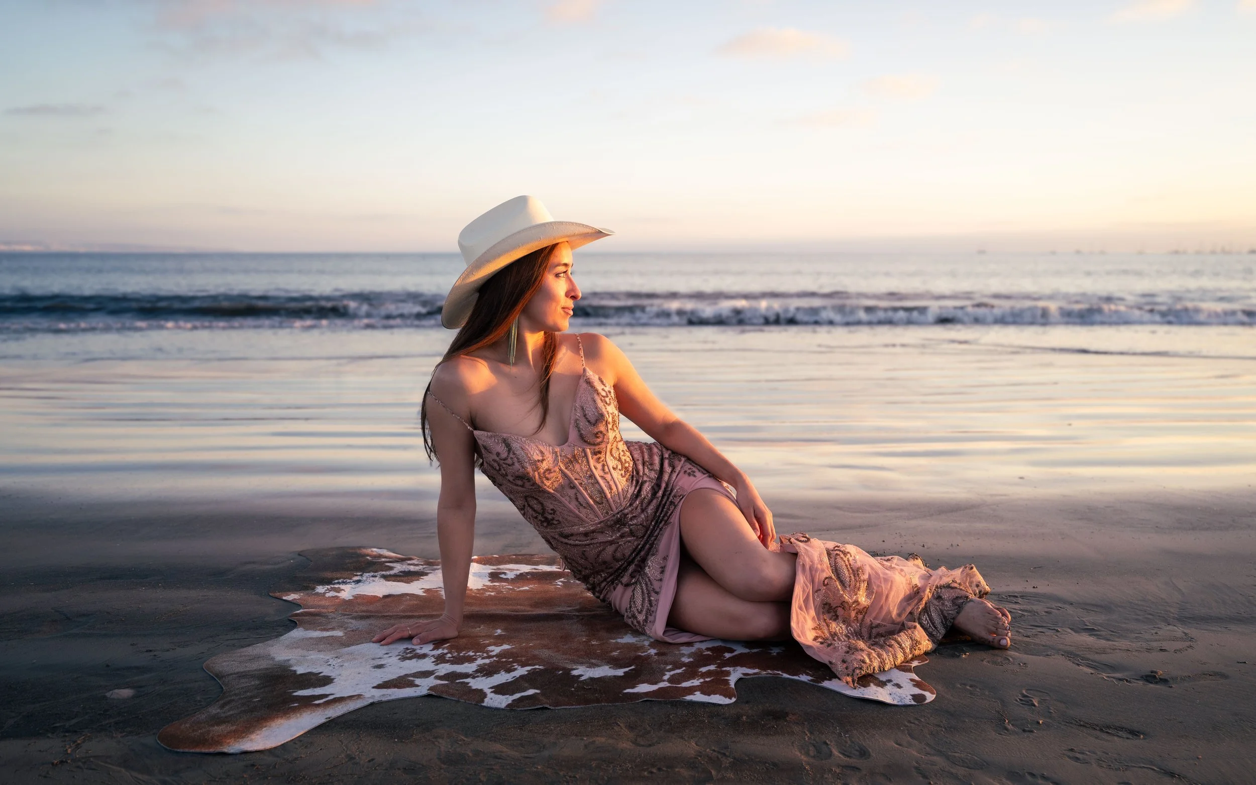 A woman in a pink dress and wide-brimmed hat sitting on the beach near the water at sunset.