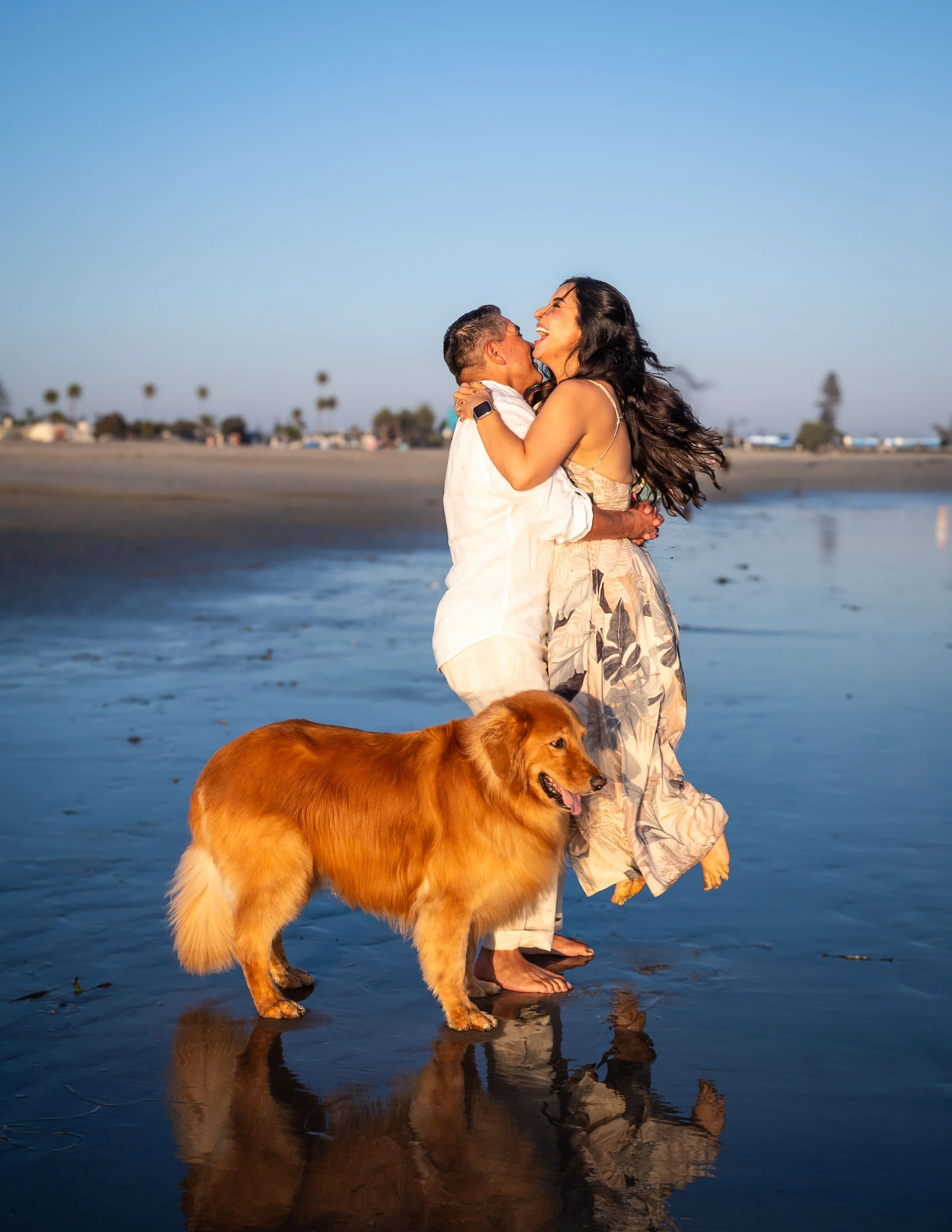 A couple is hugging and dancing on the beach with a golden retriever. The woman has long dark hair and is wearing a light dress, while the man is wearing a white shirt and shorts. There is a clear blue sky and palm trees in the background.