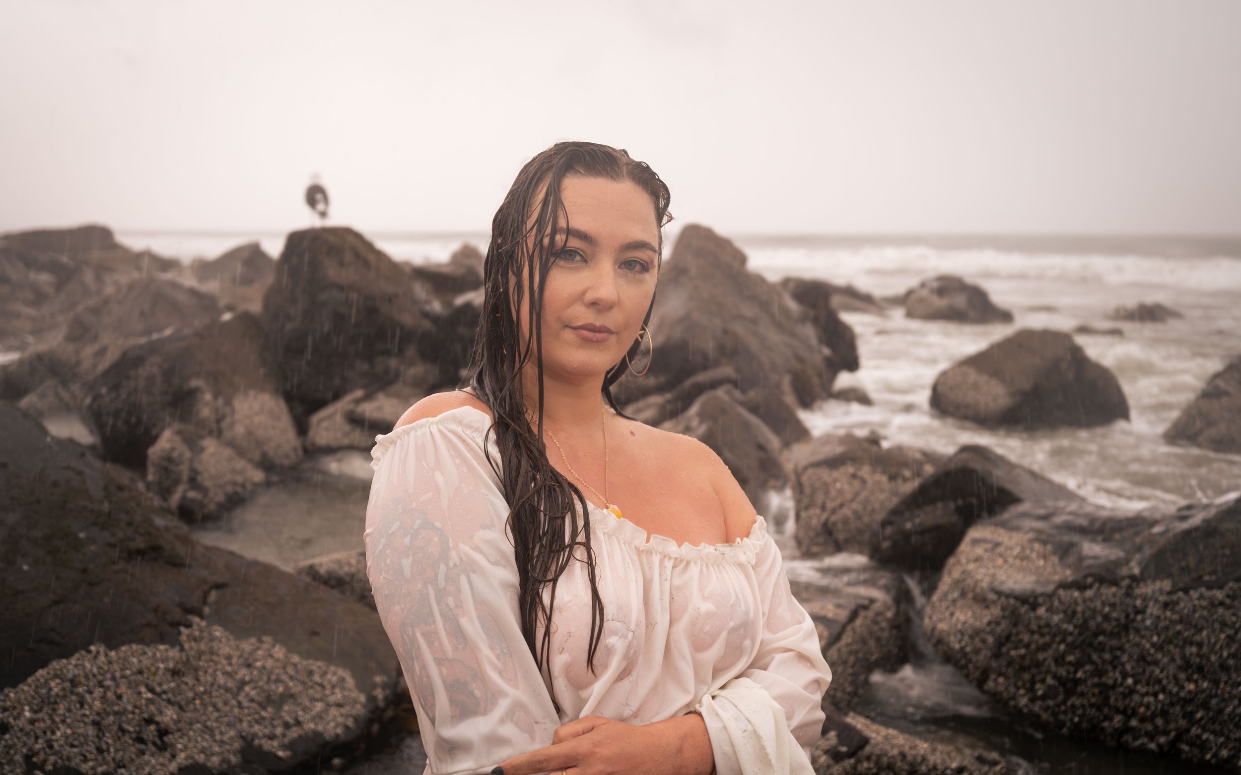 A woman with wet dark hair and blue eyes standing on a rocky beach with waves crashing behind her, wearing a white off-shoulder top.