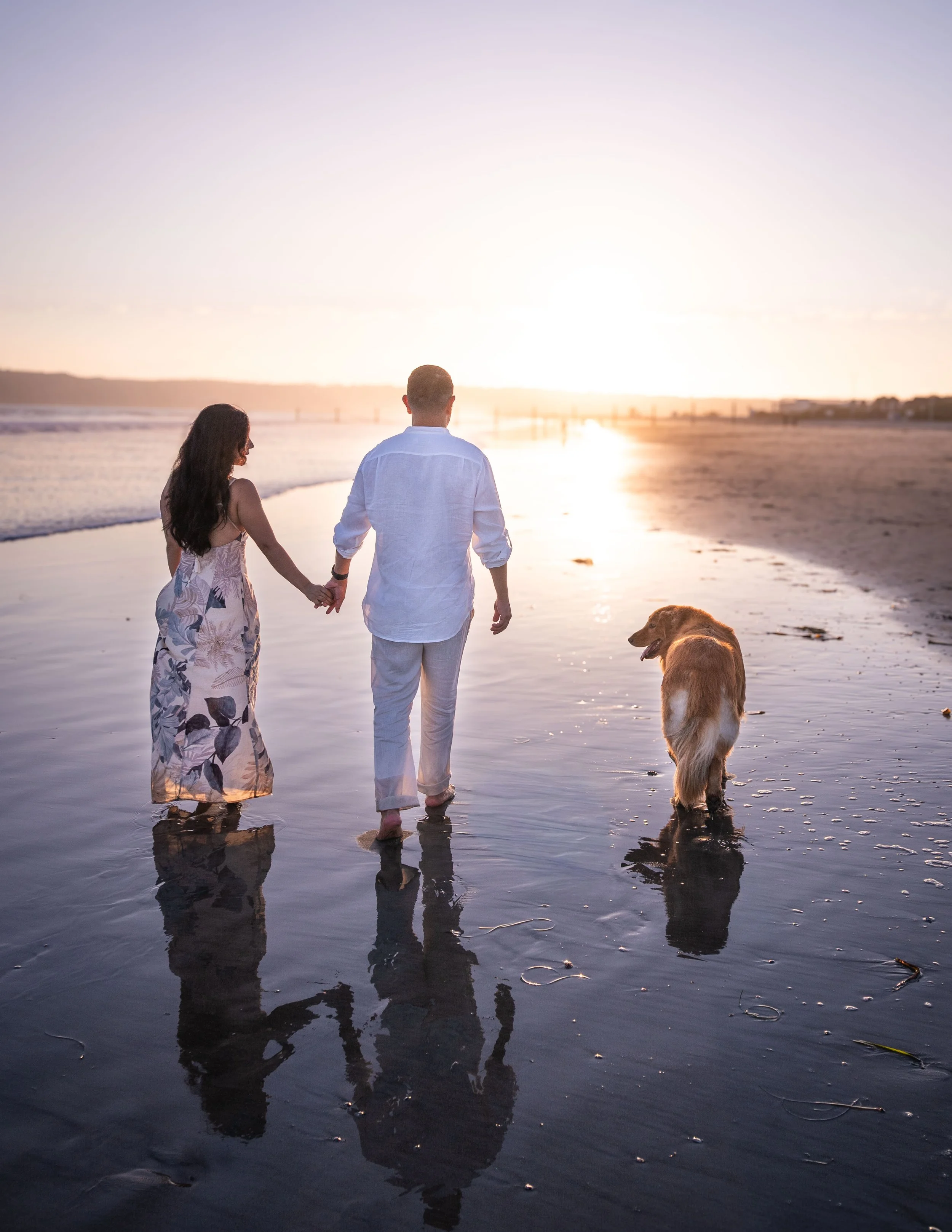 A couple holding hands walks along the beach at sunset, accompanied by a golden retriever dog. The scene reflects a calm and romantic atmosphere with the sun low on the horizon and the sky glowing softly.