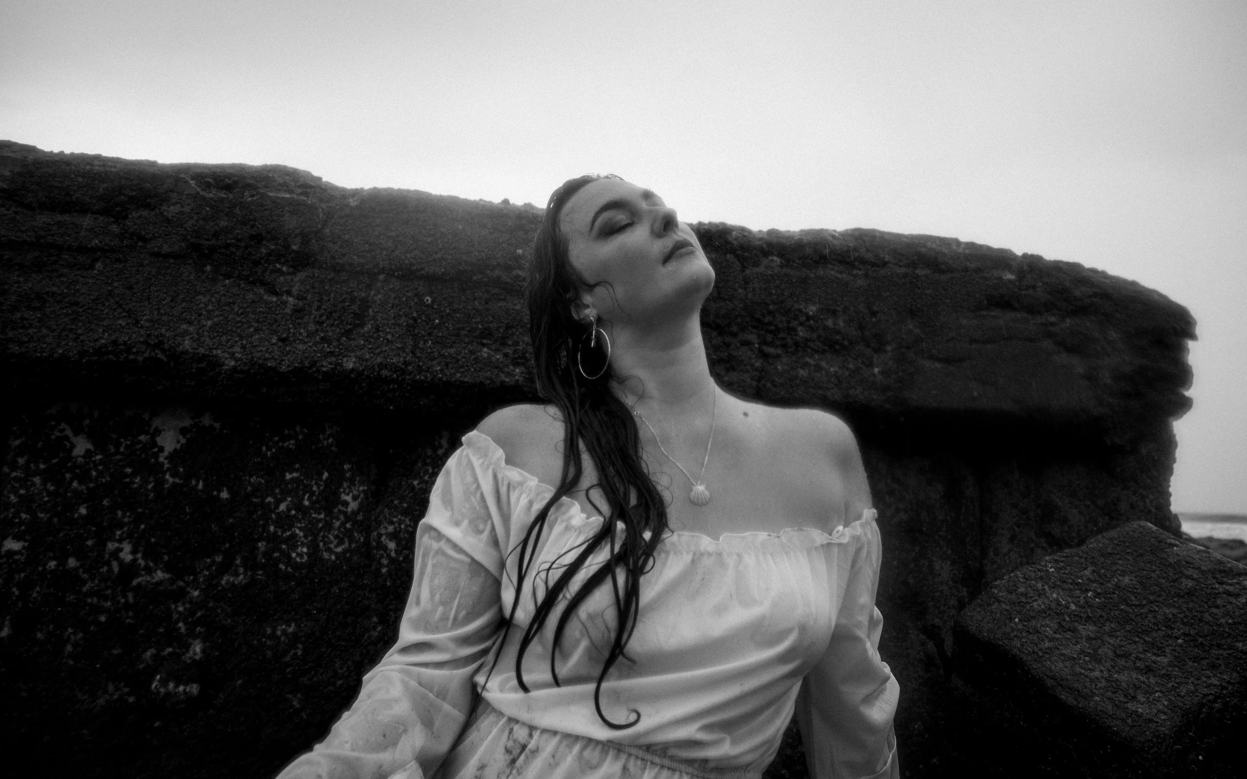 Black and white photo of a woman with wet hair in a white off-shoulder dress, standing outdoors near large rocks, with her eyes closed and head tilted back.