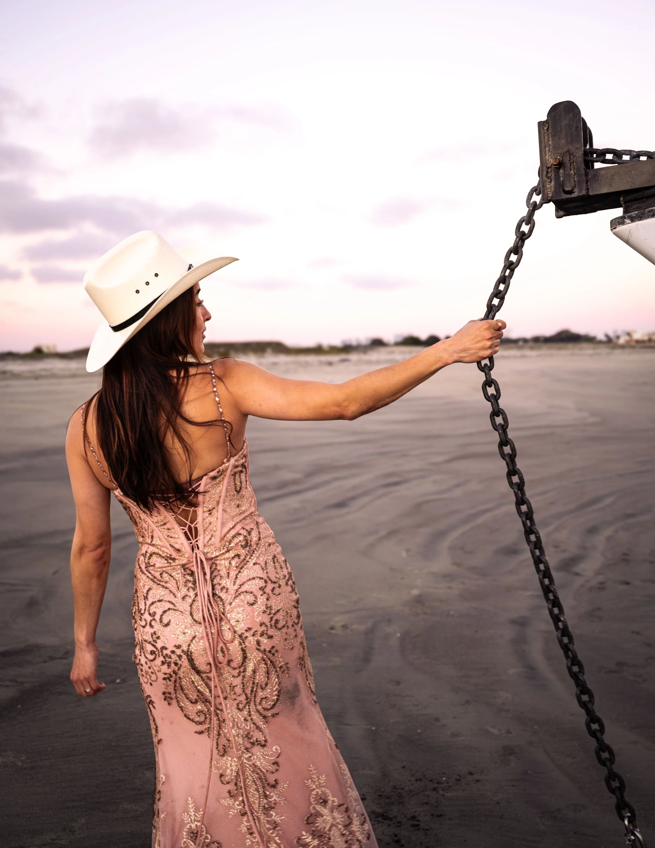A woman in a pink, embroidered gown and a white wide-brimmed hat stands on a beach holding a chain attached to a boat or dock. The sky is cloudy and the ocean is in the background.