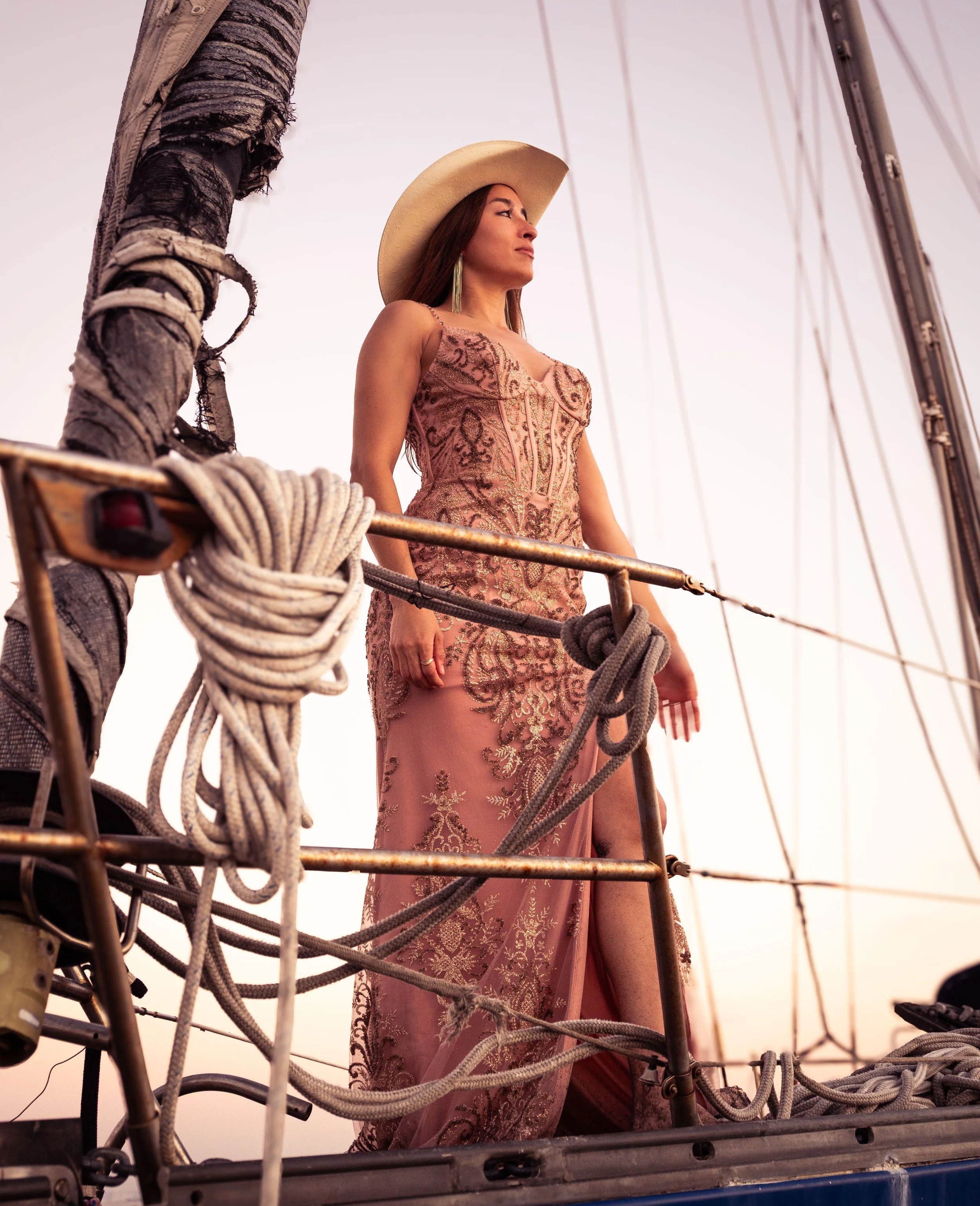 A woman in a pink, embroidered dress and wide-brimmed hat standing on a boat, looking into the distance at sunset.