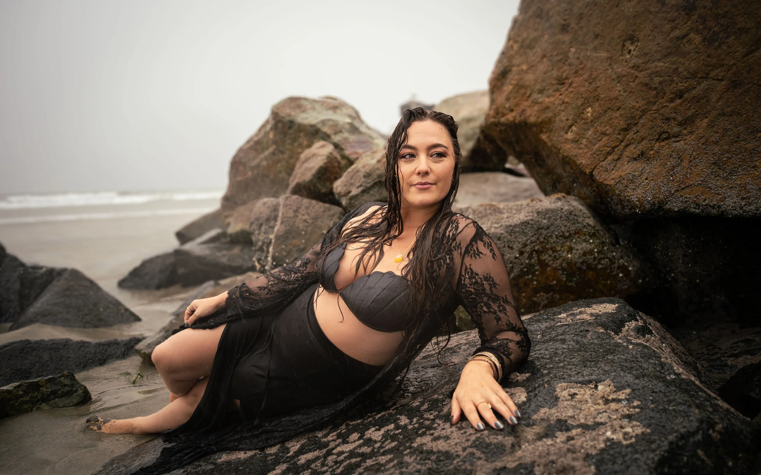 Woman with wet hair lounging on rocks at the beach, wearing black lace cover-up and black swimsuit, with ocean waves in the background.