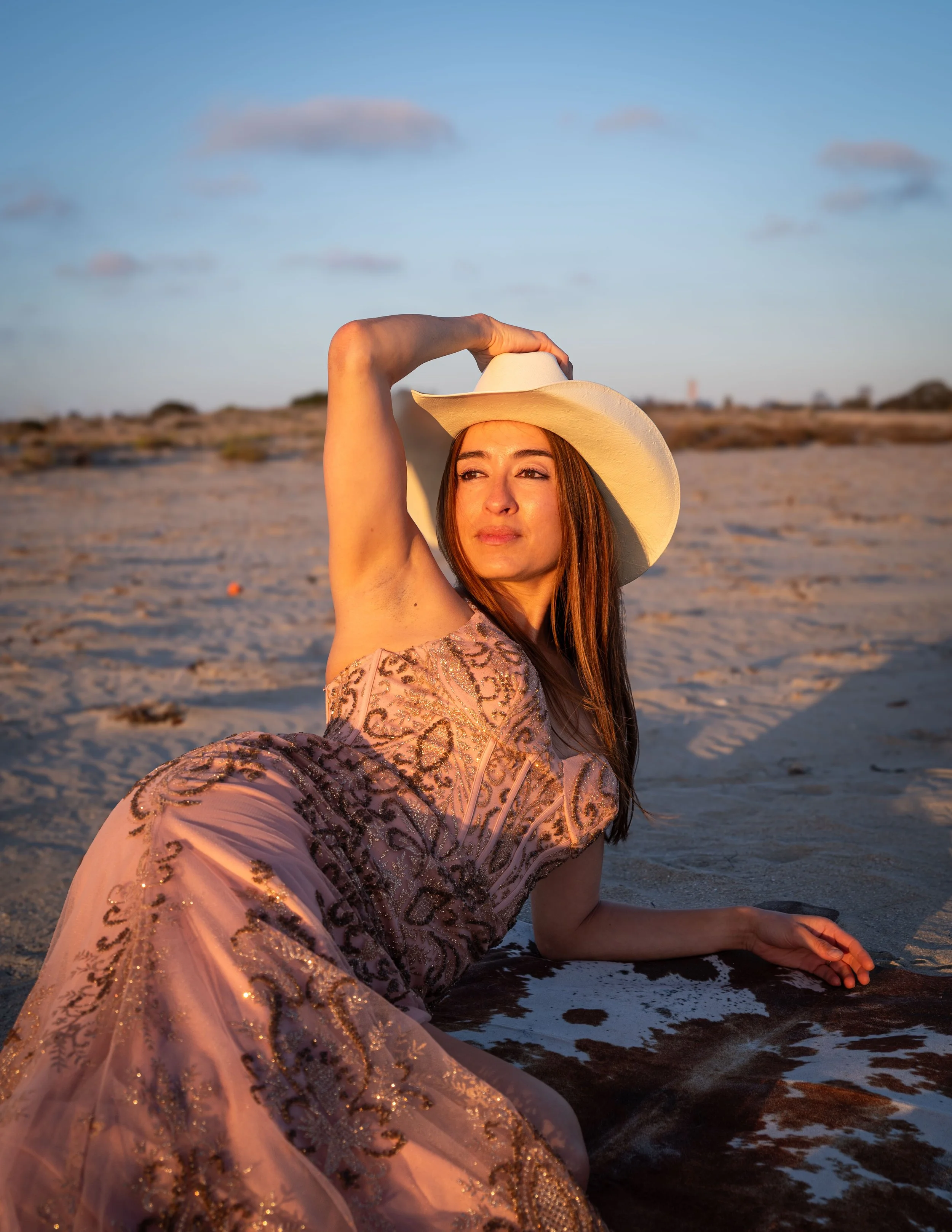 A woman sitting on the beach at sunset, wearing a pink dress with gold embroidery and a white cowboy hat, with her arm raised and holding the hat. The ocean and sky are in the background, with some clouds and a calm sea.