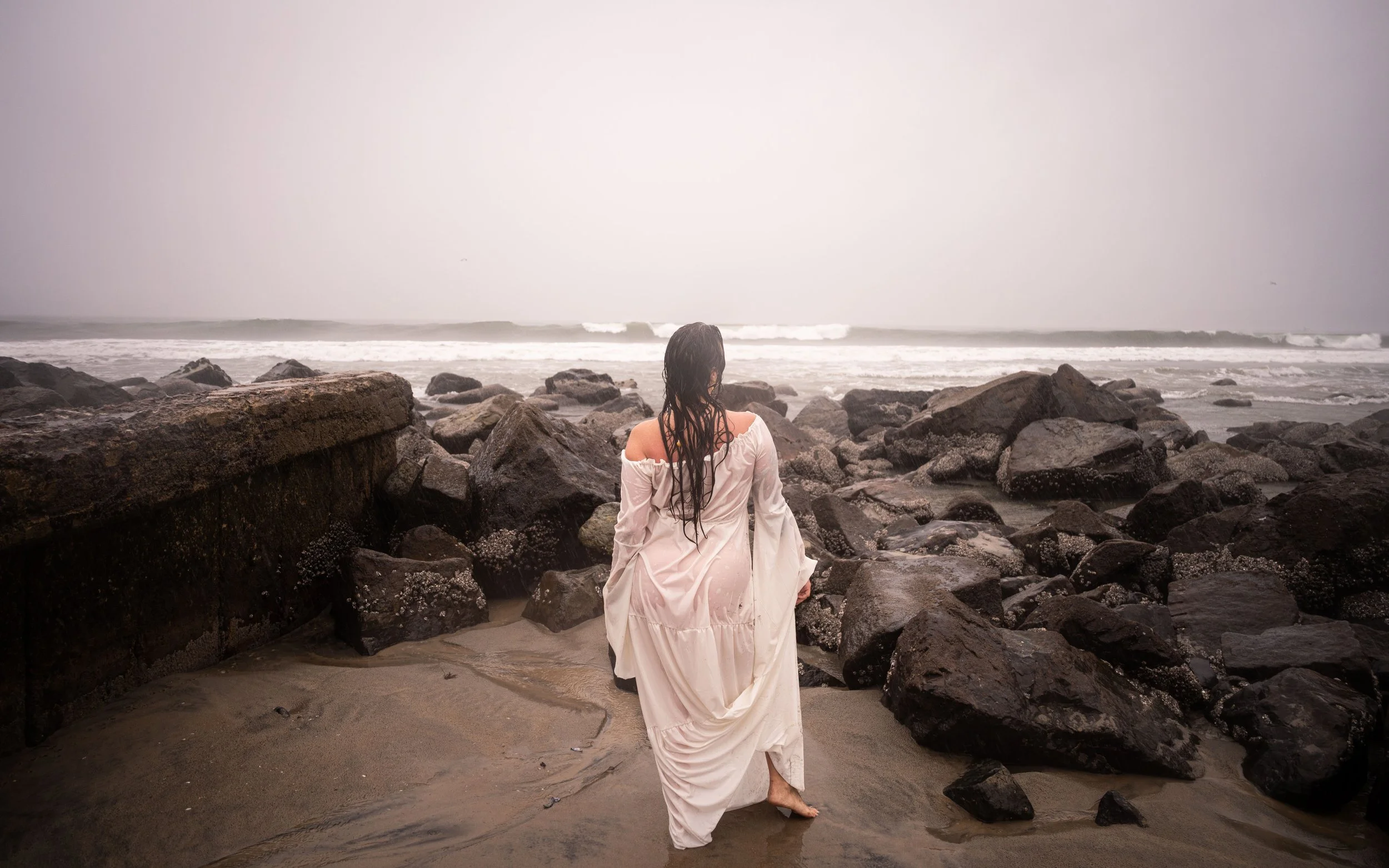 A woman with long, wet dark hair in a white dress kneeling on rocks at a beach, looking toward the ocean on a cloudy day.