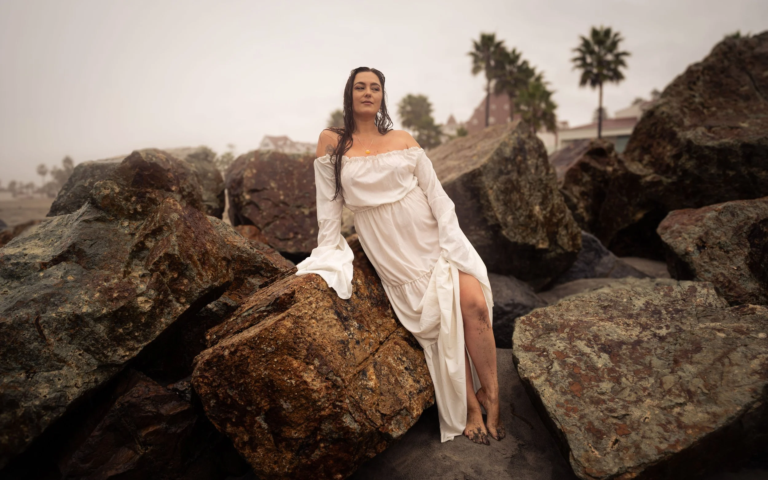 A woman in a white off-shoulder dress sitting among large rocks on a beach with palm trees and houses in the background during cloudy weather.