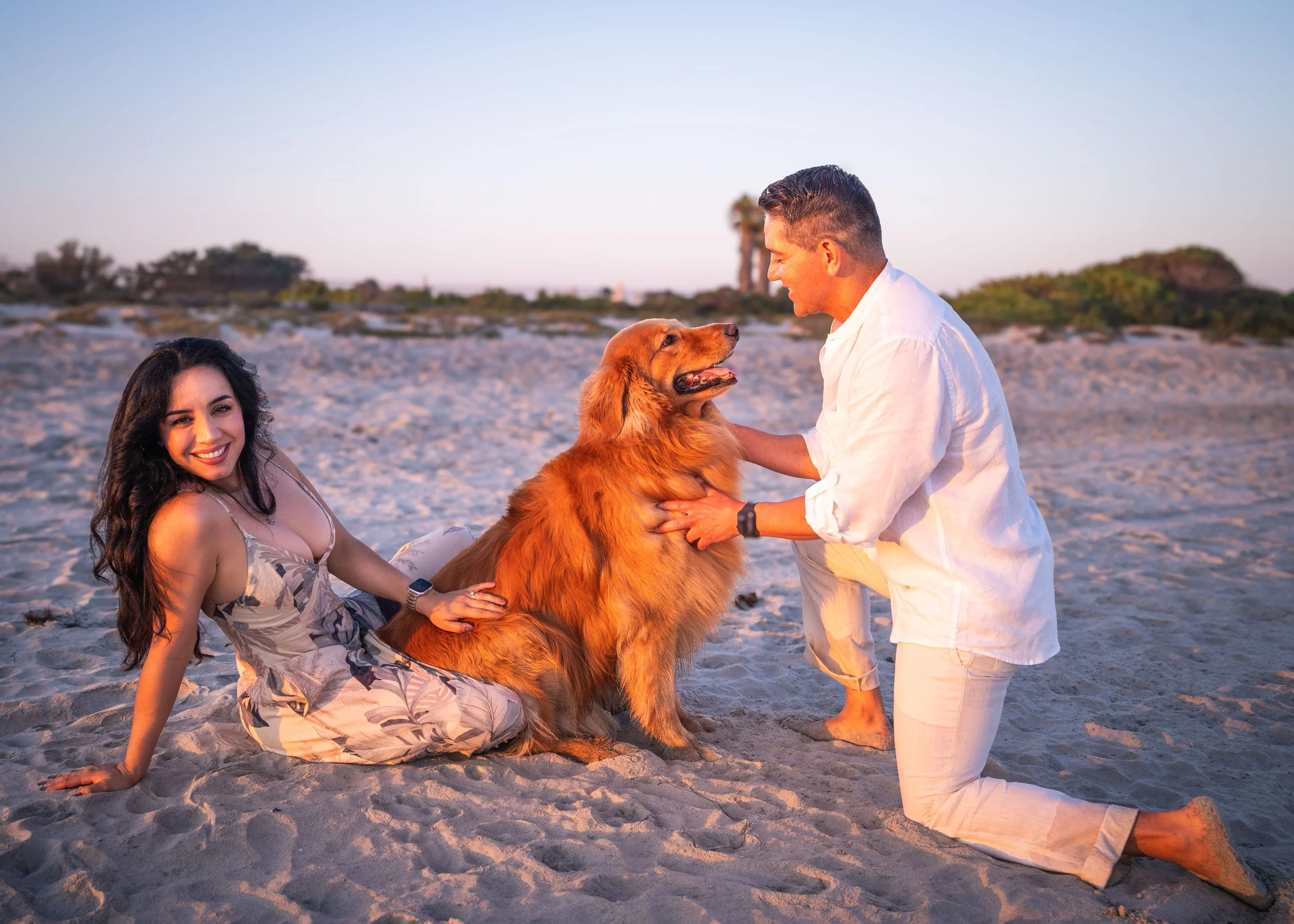 A couple with a golden retriever on the beach during sunset. The woman is squatting on the sand, smiling at the camera, and the man is kneeling, holding the dog, smiling at the dog. The background shows a clear sky and some vegetation.