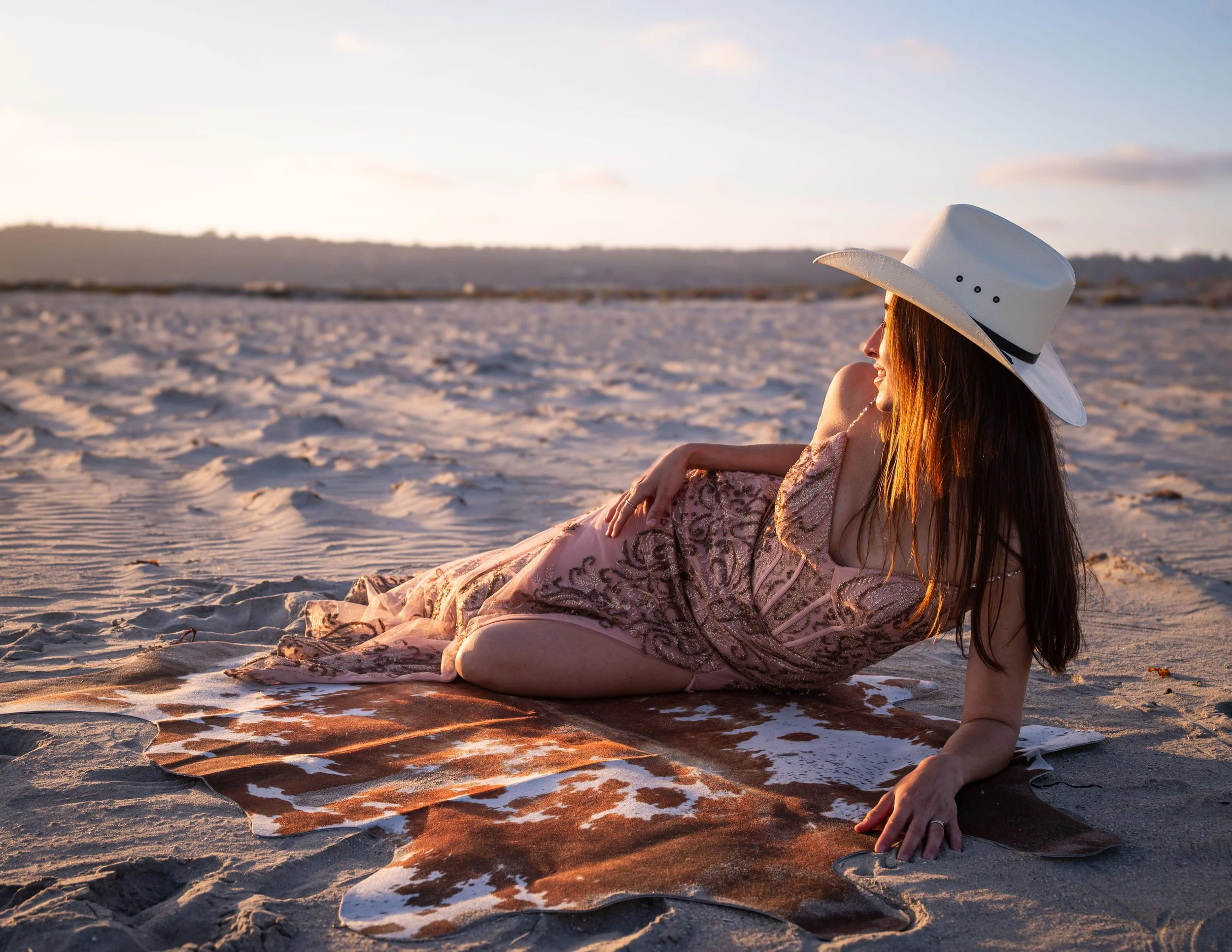 A woman in a floral dress and wide-brimmed hat sits on a sandy beach during sunset, with her side profile visible as she relaxes and smiles.