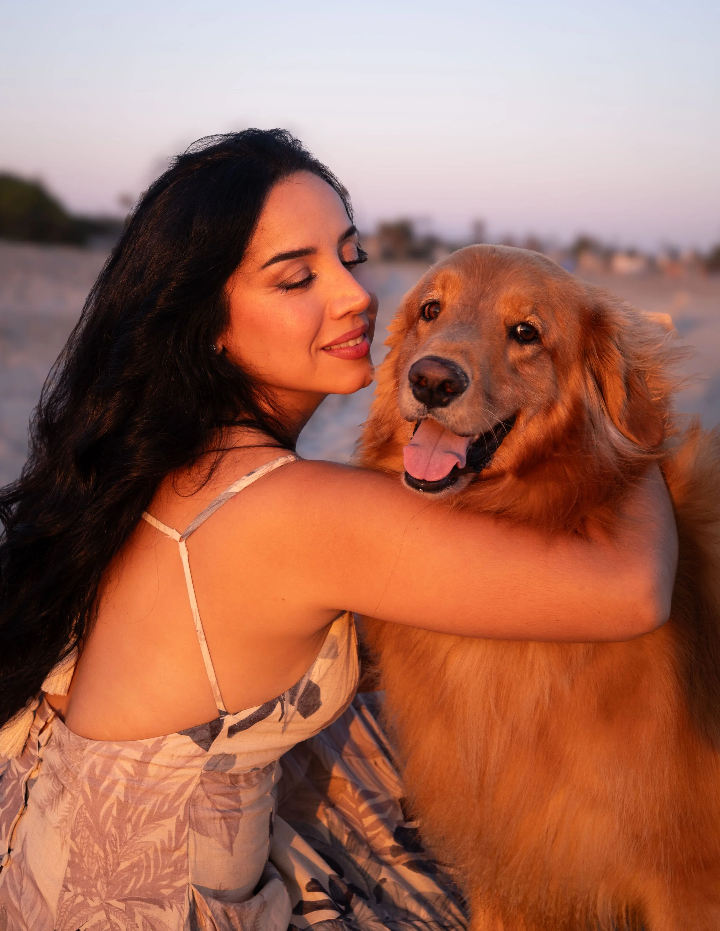 A woman with long dark hair and closed eyes hugging a golden retriever dog outdoors during sunset.