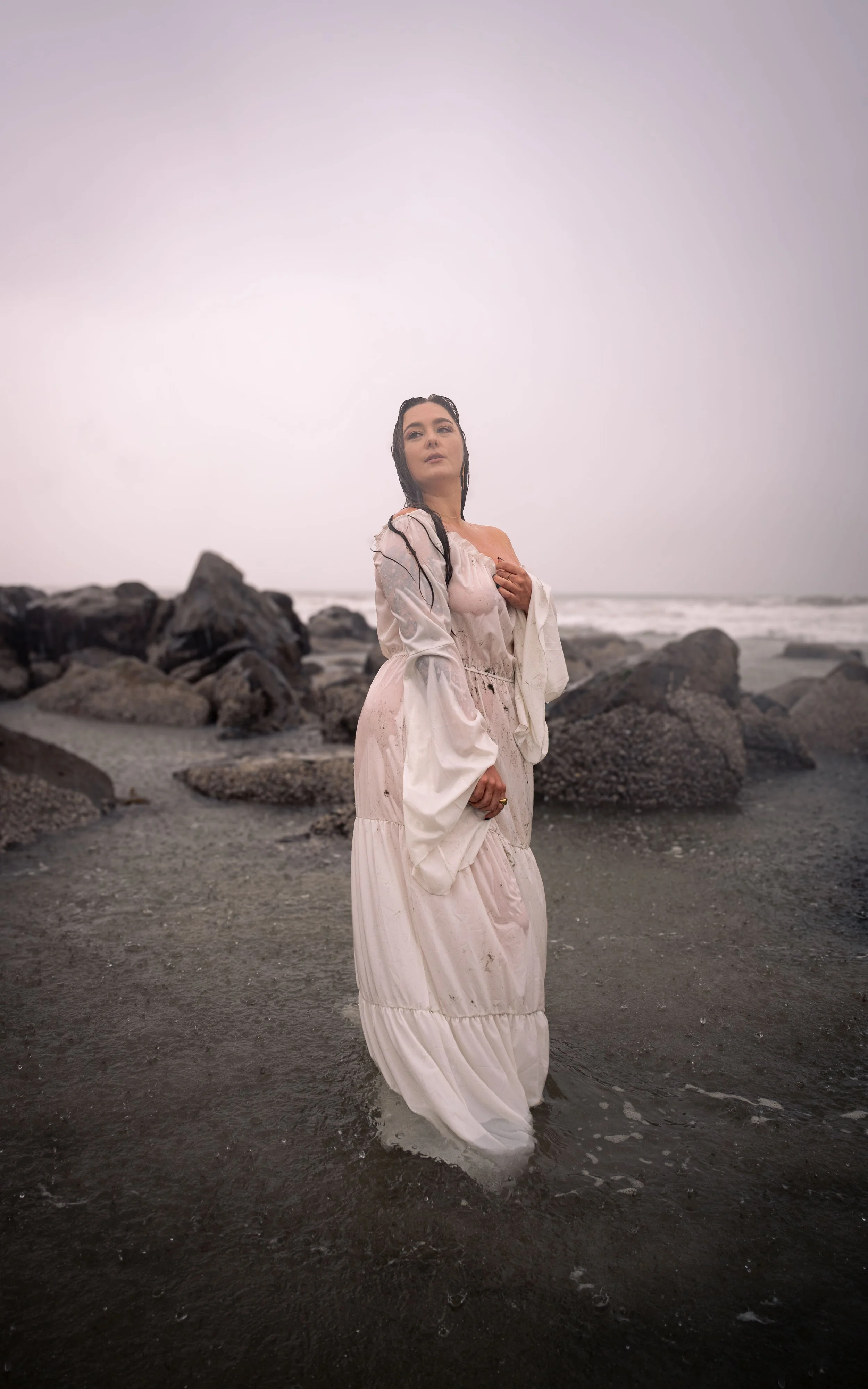 A woman in a long, flowing, white dress standing at the edge of the ocean on a cloudy day, with rocks and waves in the background.