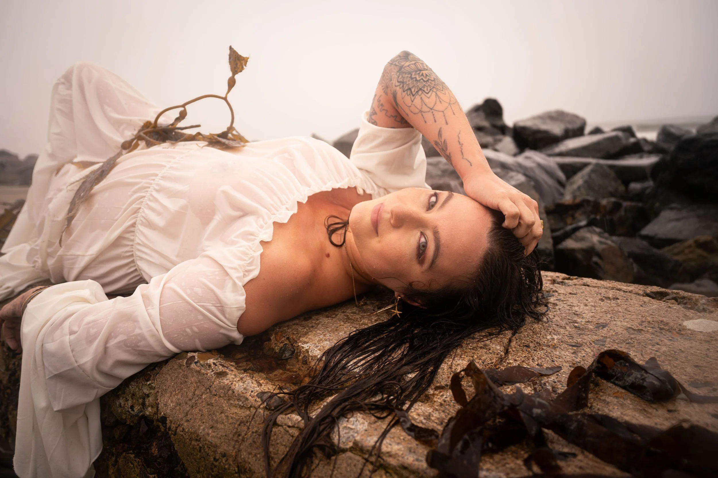 A woman lying on a rocky beach, wearing a white dress, with dark hair spread out, and looking at the camera. There are rocks and seaweed in the background.