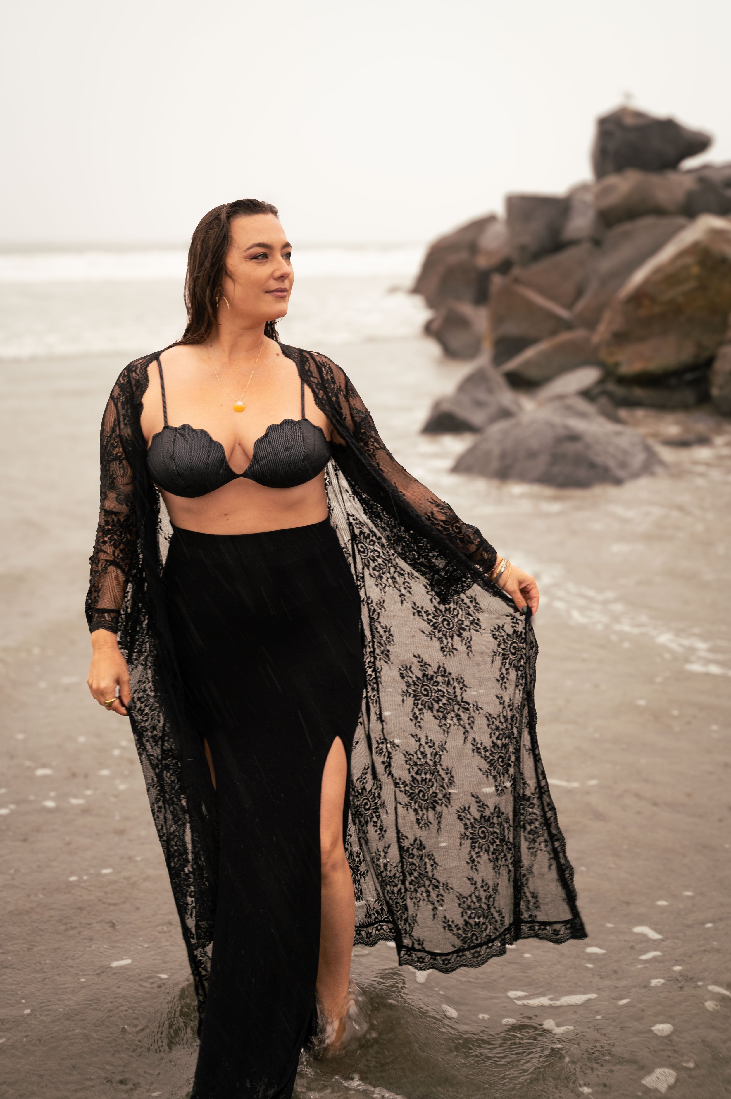 A woman in a black lace dress with a slit walking on the beach near rocks, with wet hair, and cloudy overcast sky.