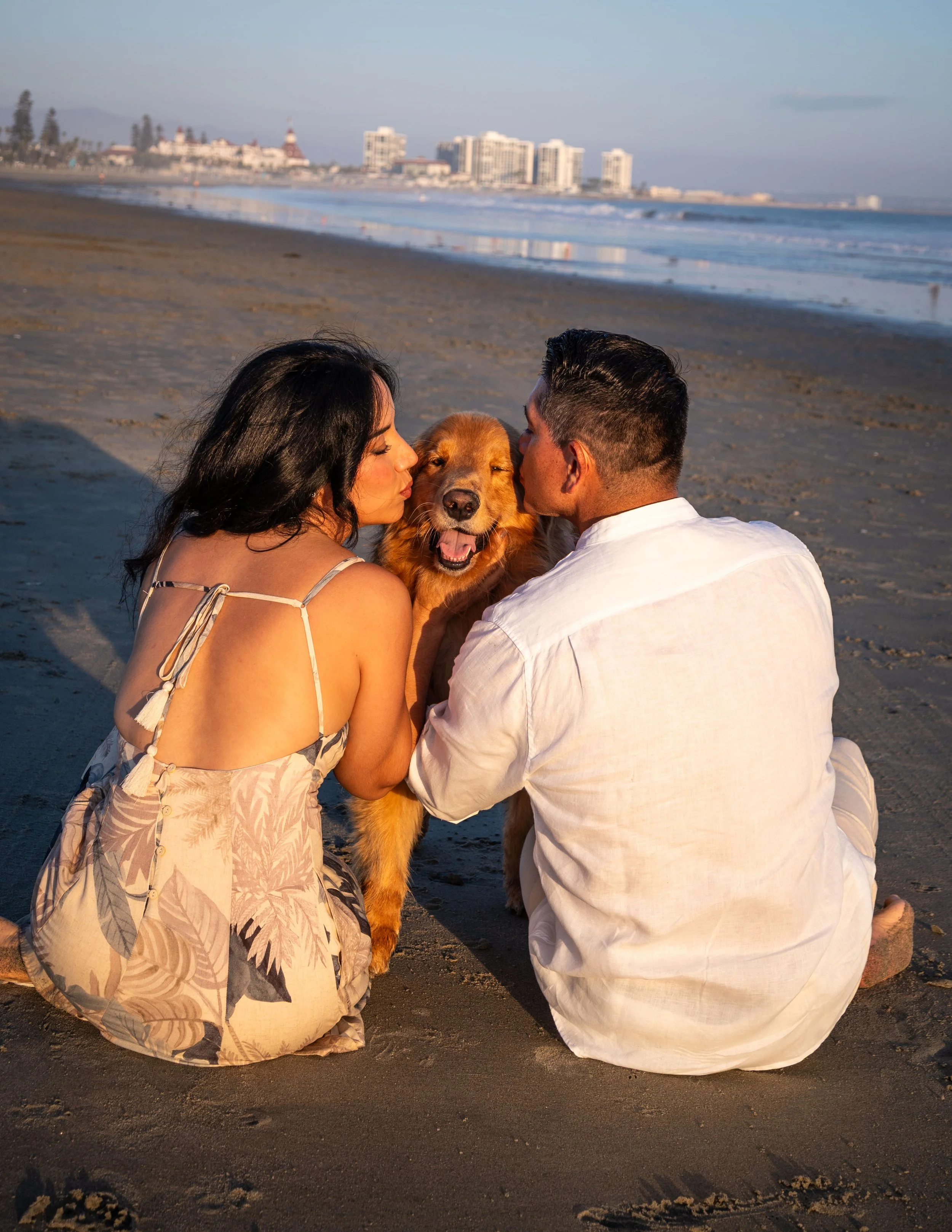 A couple sitting on the beach with their dog at sunset, facing each other and giving kisses to the dog in the middle. The city skyline is visible in the background.