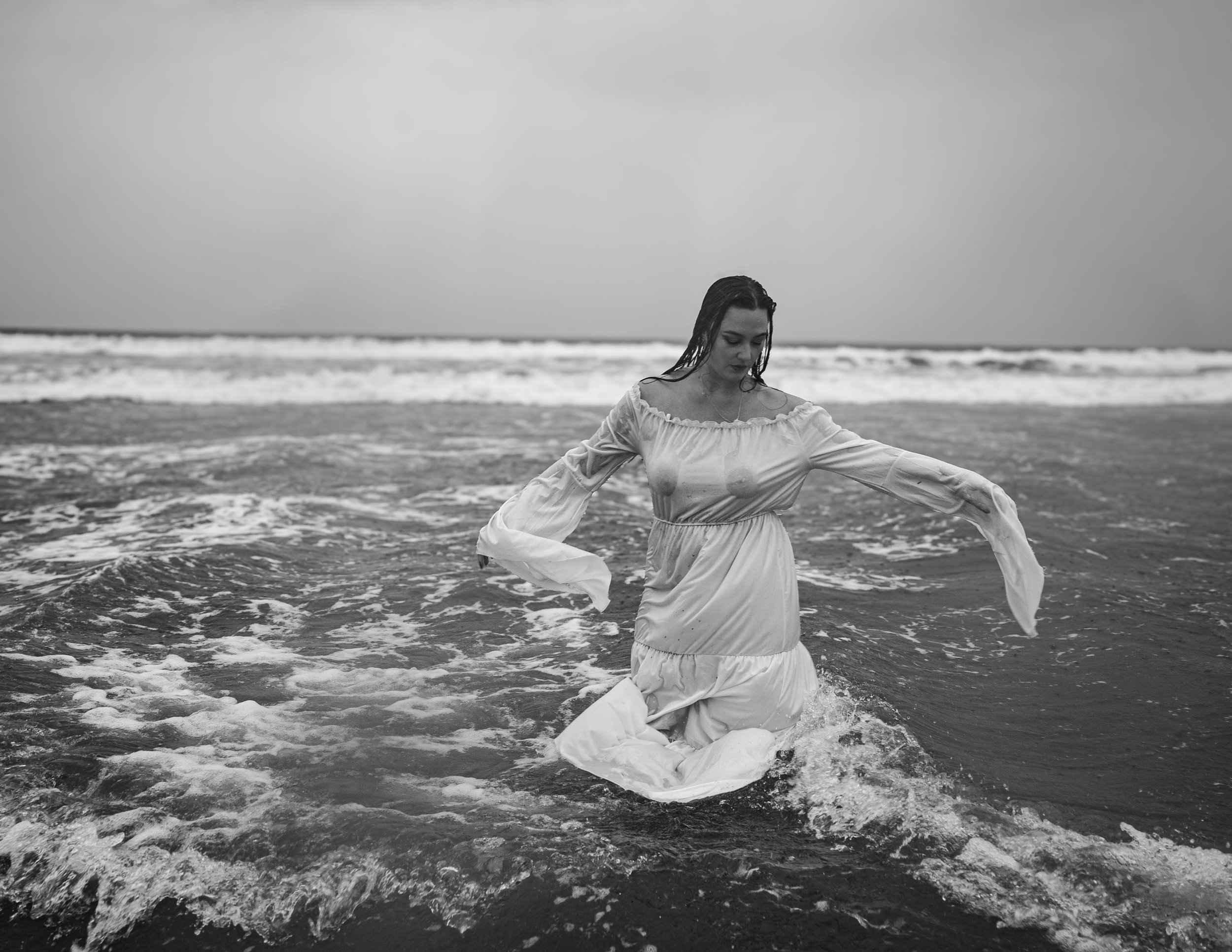 A woman with wet hair wearing a long dress stands in the ocean waves, looking down with her arms extended. The sky is cloudy, and the scene is black and white.