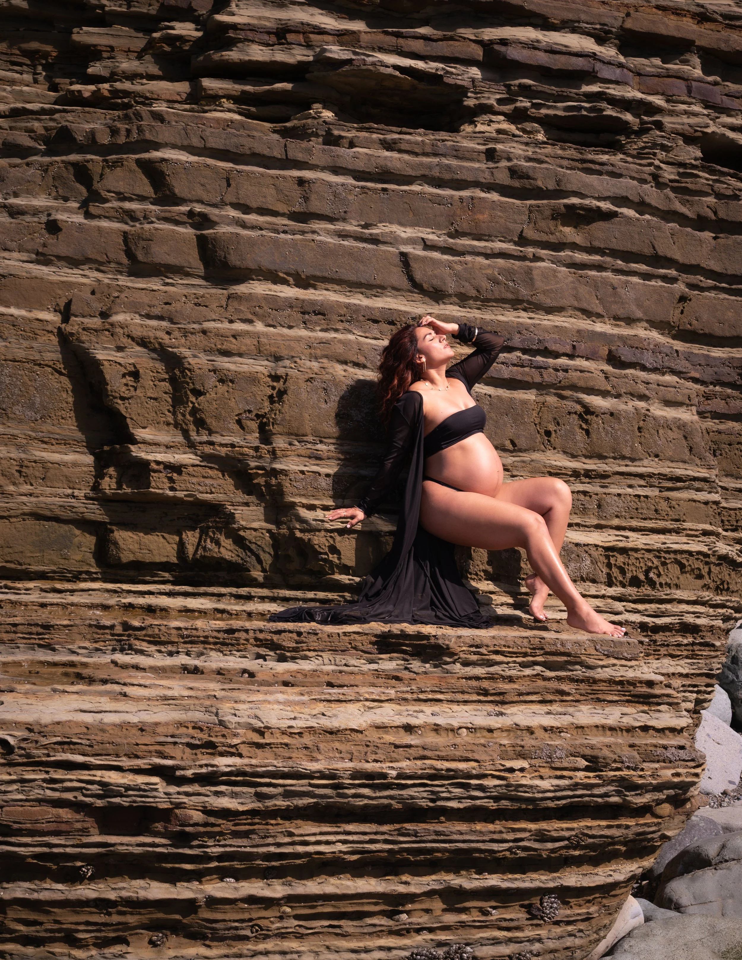 A pregnant woman in black attire posing against a layered rock wall at the beach, with her hand on her head and eyes closed, enjoying the sunlight.