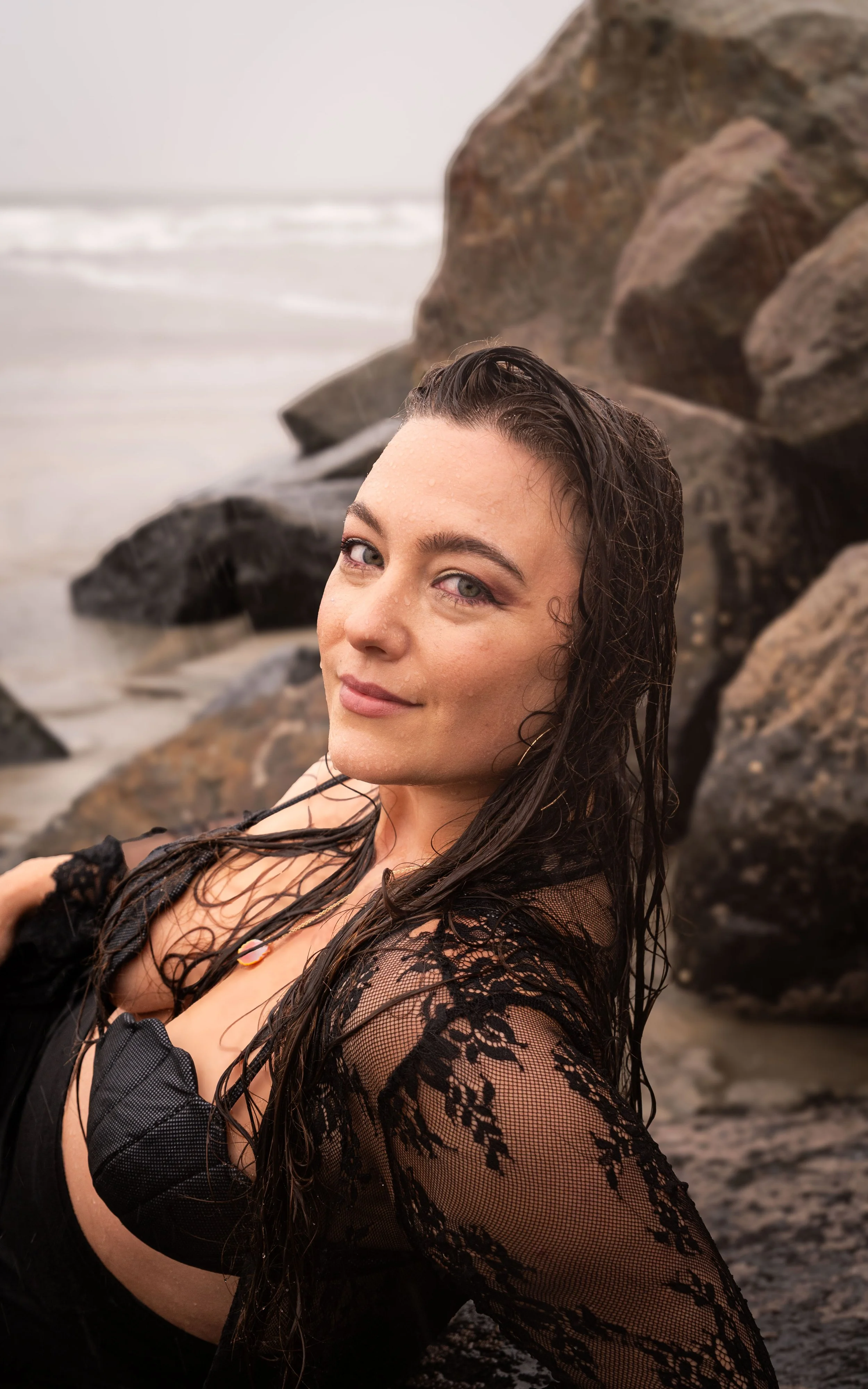 A woman with wet hair and a lace black top posing near rocks on the beach with the ocean in the background.