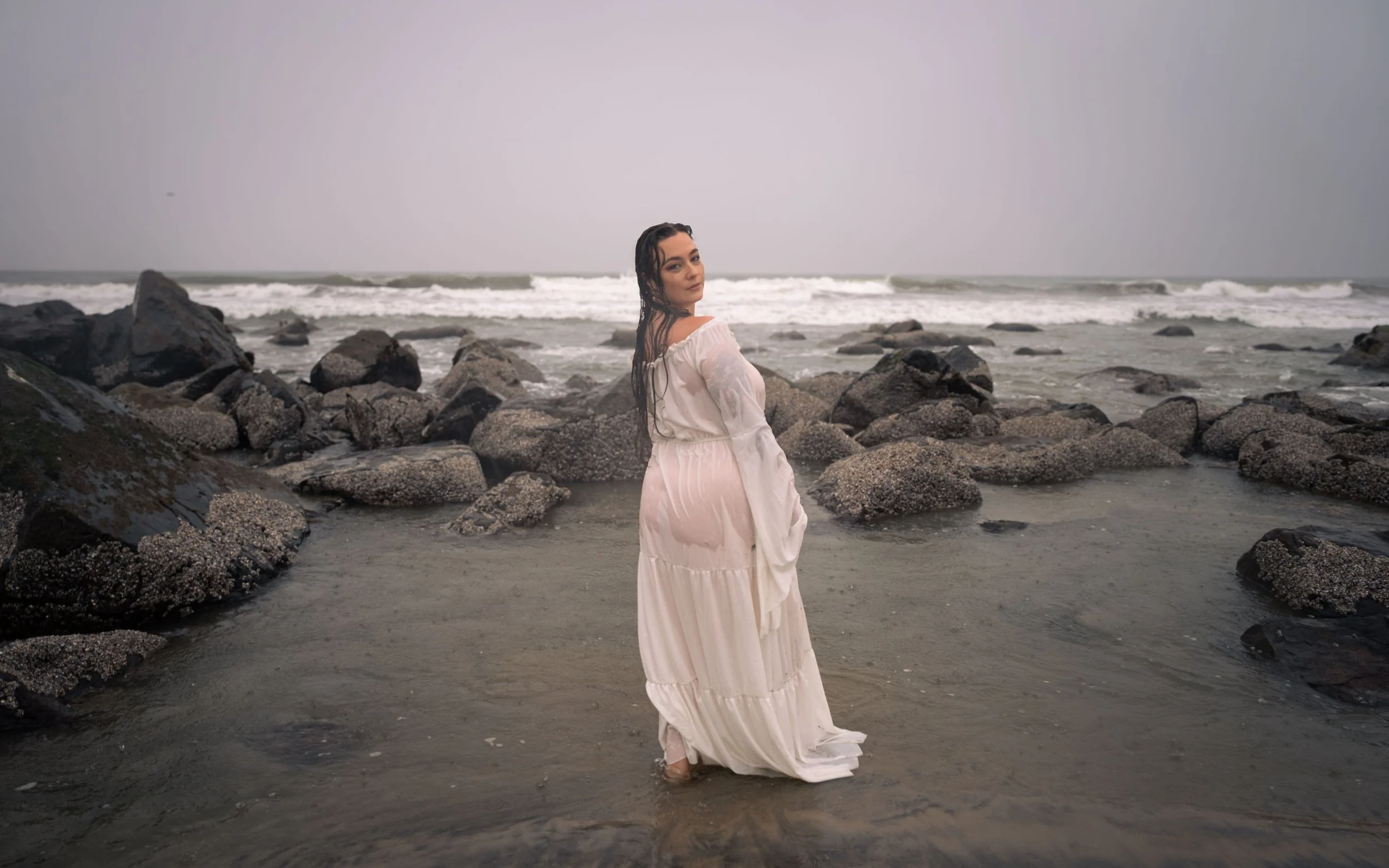 A woman in a long, light-colored dress standing in shallow water at the beach, with rocks in the foreground and waves in the background under a cloudy sky.