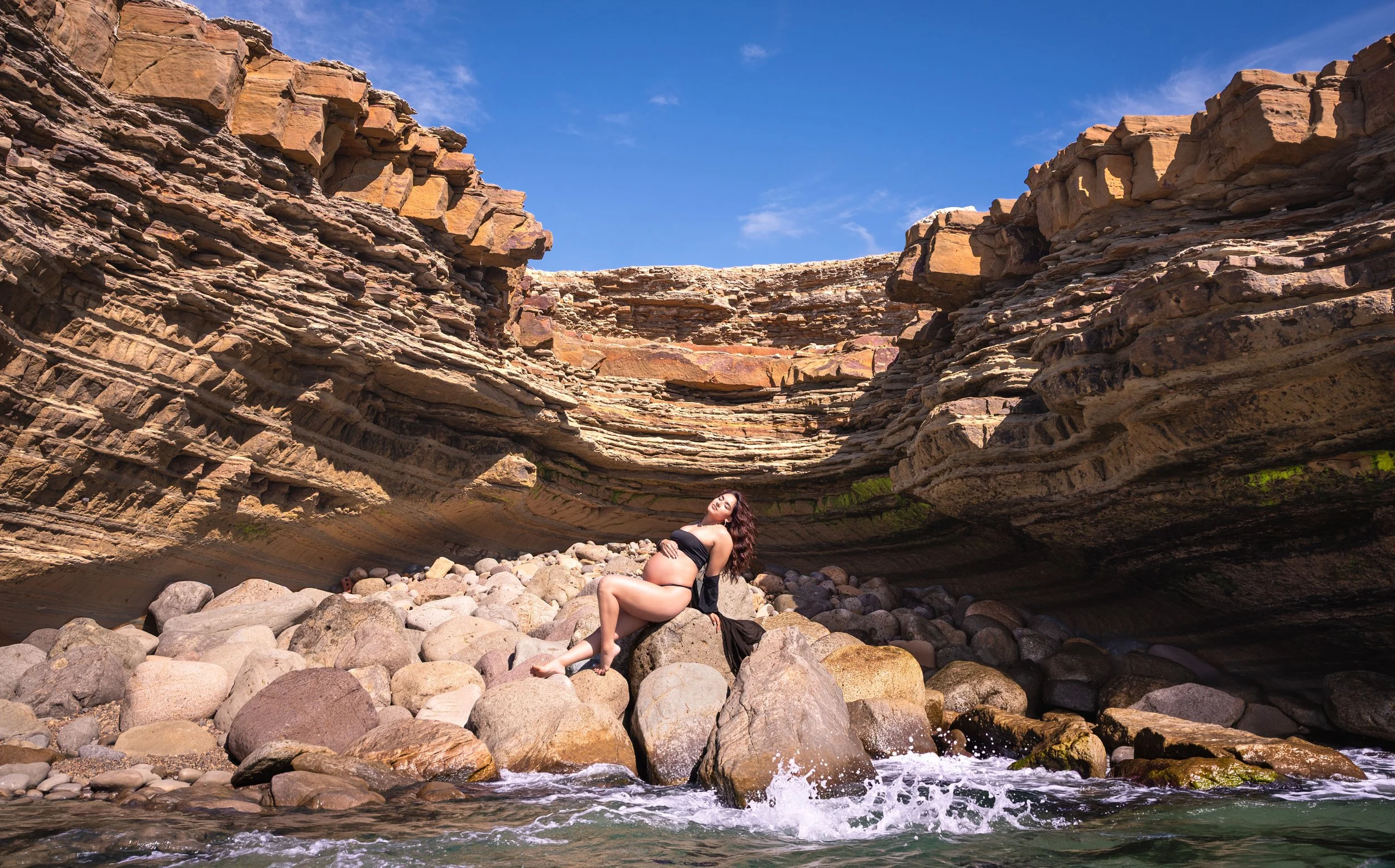A pregnant woman in a black dress sitting on rocks inside a canyon with layered rock formations and a blue sky with some clouds above.