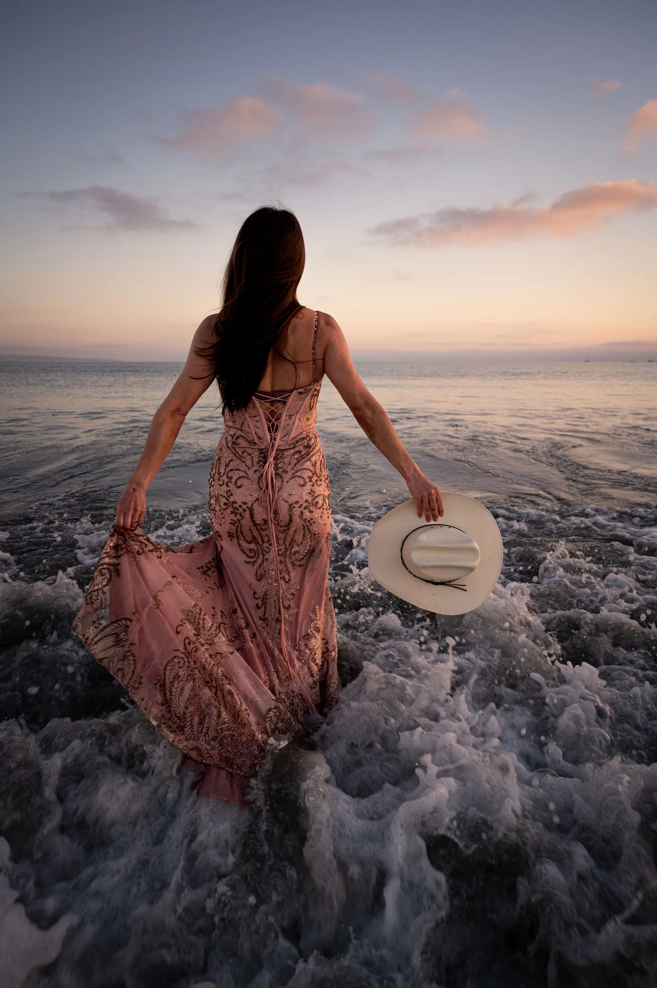 A woman in a pink, embellished dress holding a white hat stands in shallow ocean water at sunset, facing the horizon with her back to the viewer, and long hair flowing.