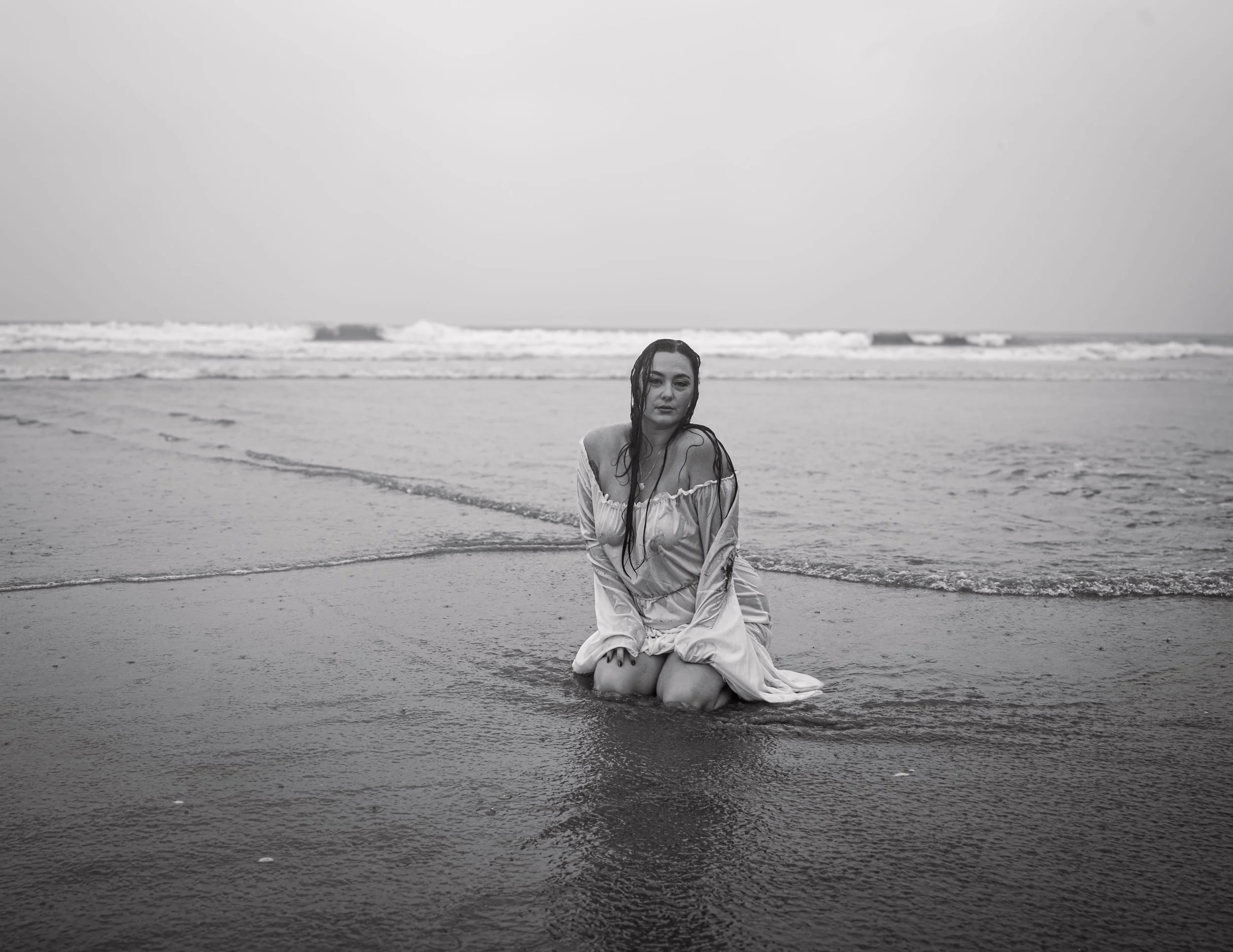 Black and white photo of a woman with wet hair sitting on the beach with ocean waves in the background.