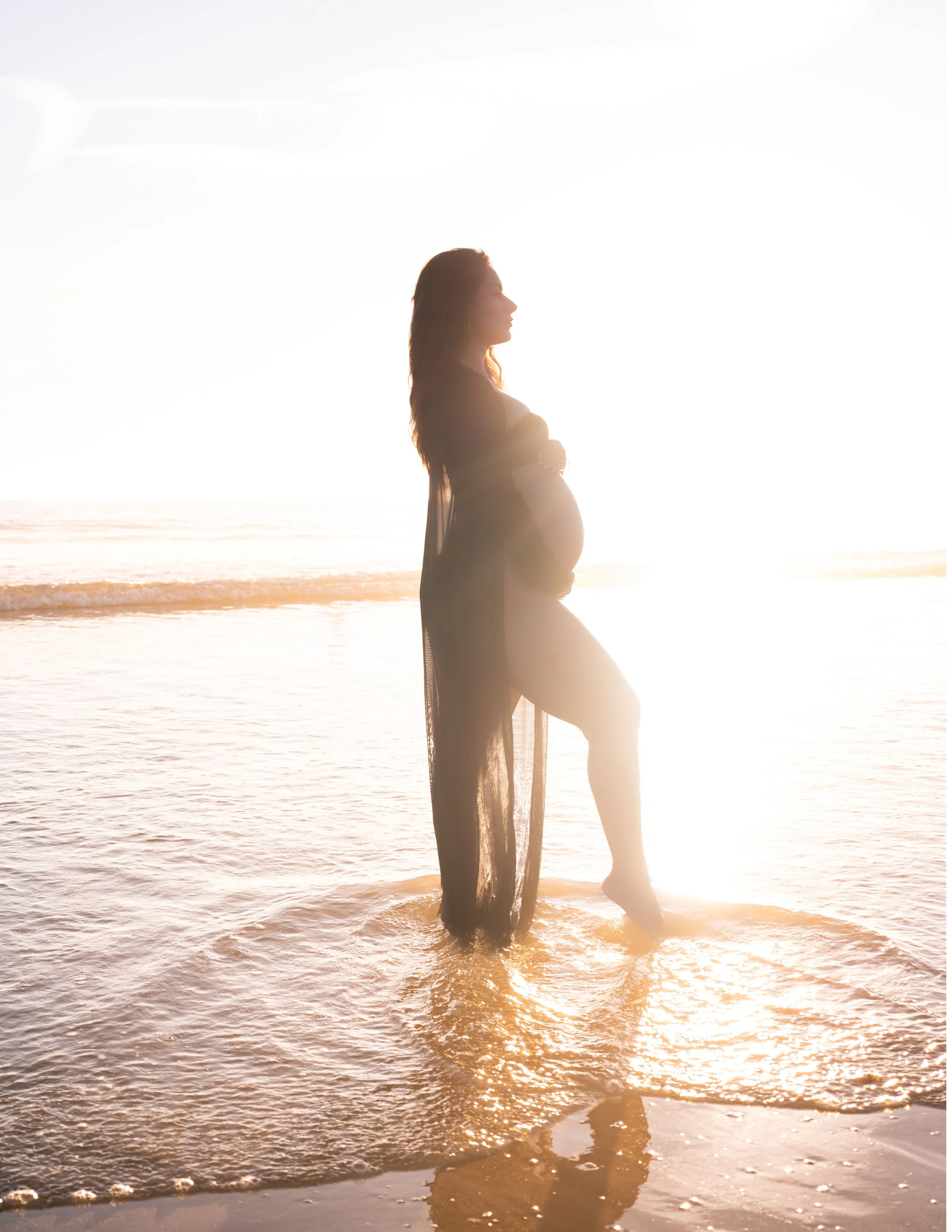 A pregnant woman standing in shallow water at the beach during sunset, with her arms crossed over her belly, and her long hair flowing down her shoulders.