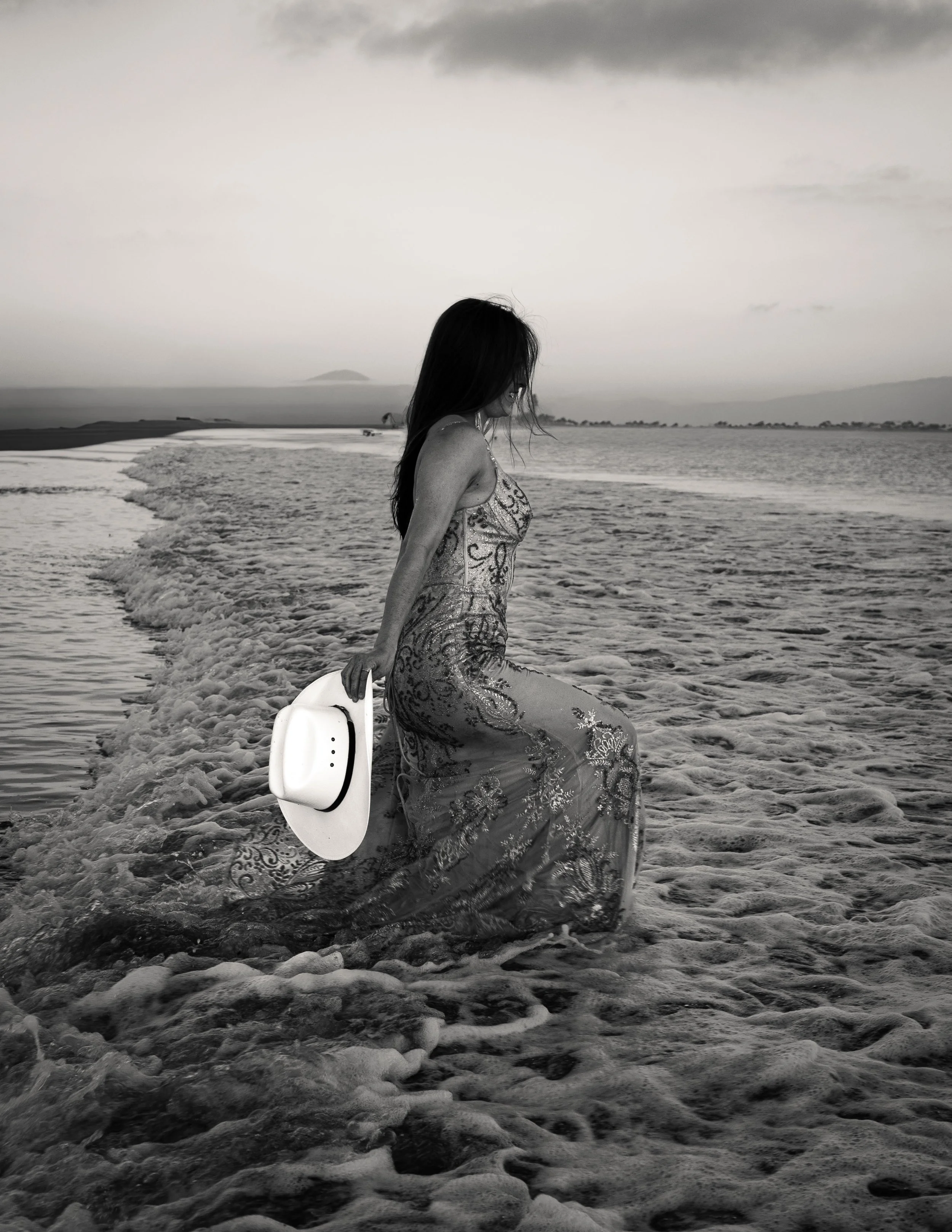 A woman in a patterned dress sitting on the wet sand of a beach, holding a white hat with both hands, with shoreline and ocean waves behind her, under a cloudy sky.