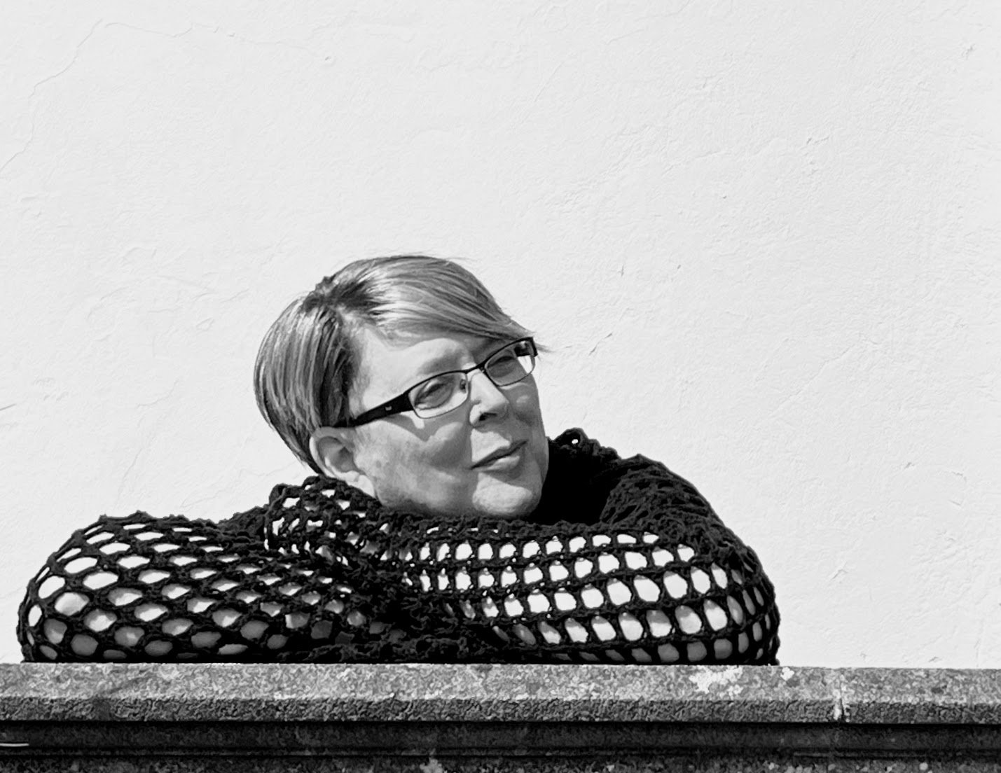 A black and white photo of Dr Char Goodwin who is leaning on their folded arms and looking to the right hand side. Char is wearing a woollen netted hoodie