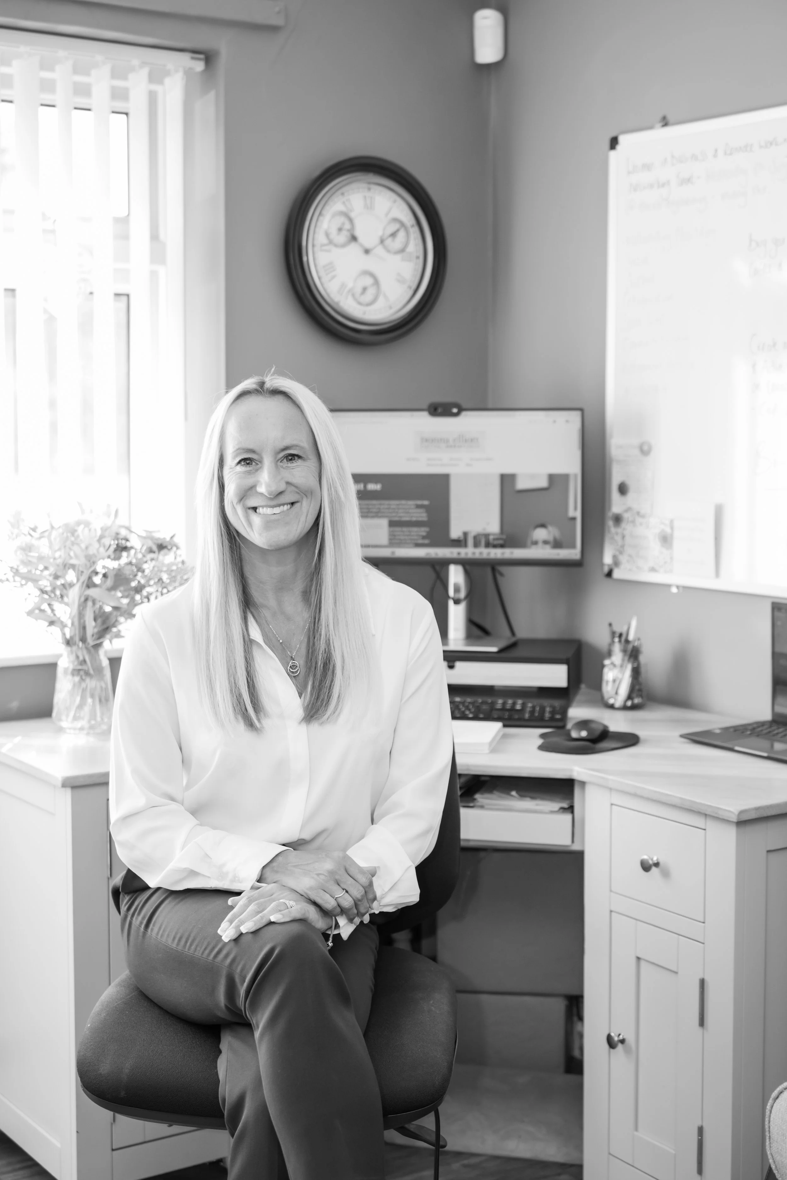 A black and white photo of Donna who is sat facing the camera with a computer on a desk behind her. She is smiling, with her hands together on her lap, and wears a white shirt and trousers