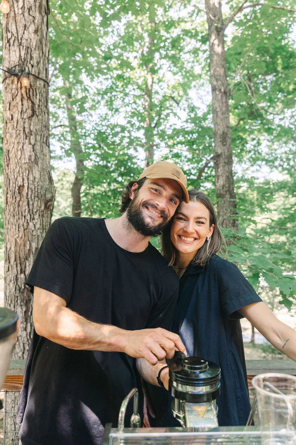 A smiling man with a beard and a woman with shoulder-length hair smiling, both standing outdoors in a wooded area, preparing coffee.
