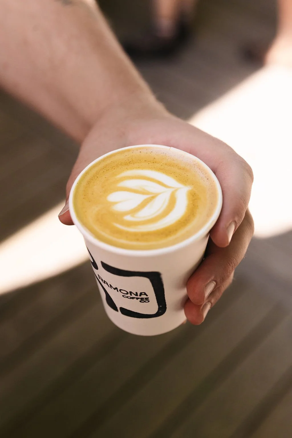 A person holding a white paper cup with the logo 'Ramona Coffee' filled with a latte featuring latte art in the shape of a rosetta.