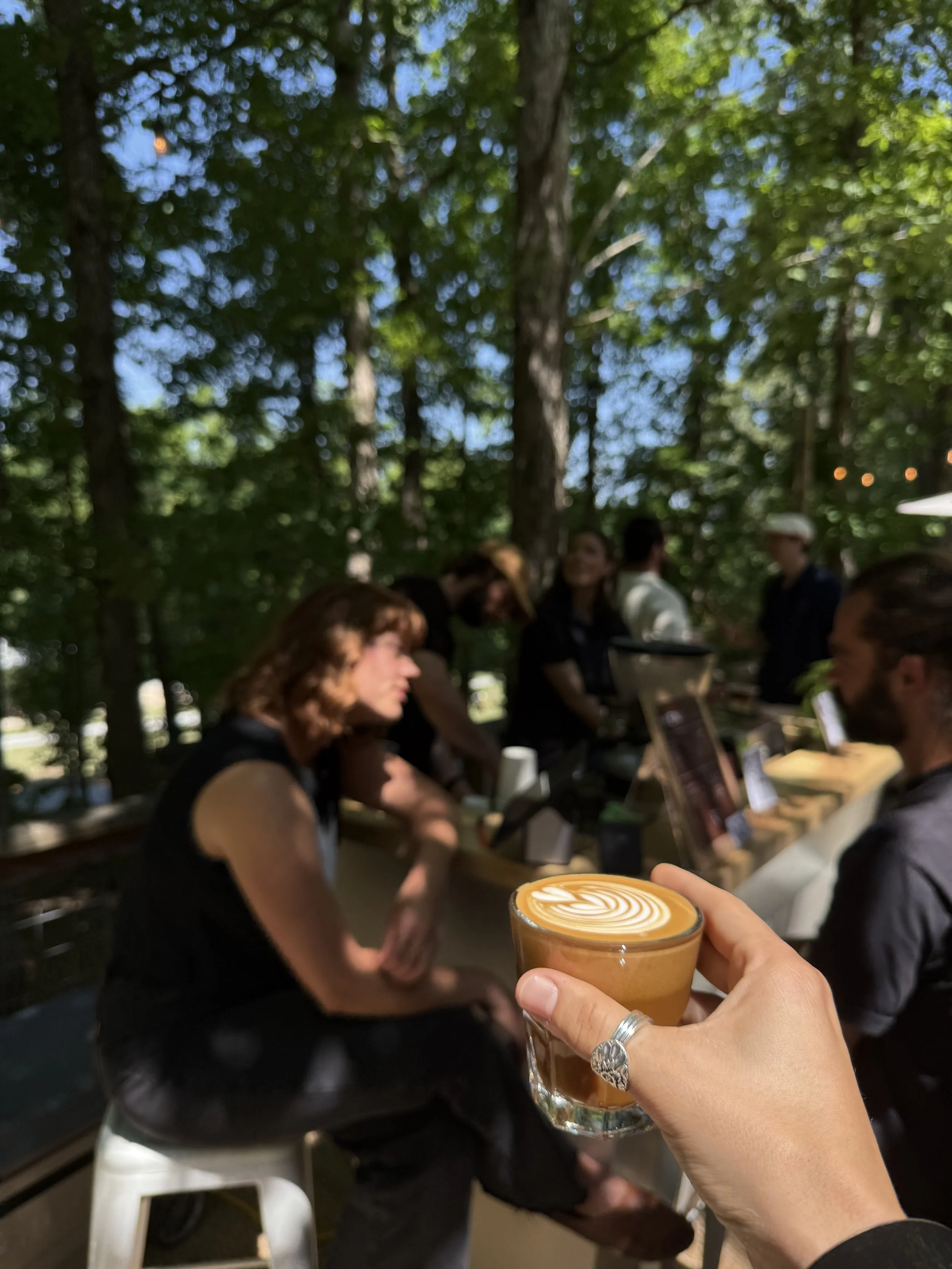 Person holding a glass of coffee with latte art in an outdoor setting with people and trees in the background.