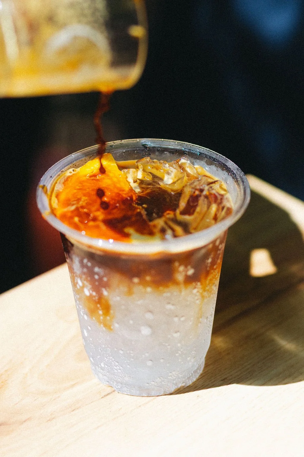 A clear plastic cup with a cold, fizzy beverage topped with crushed ice, with dark soda being poured into the cup from a bottle.